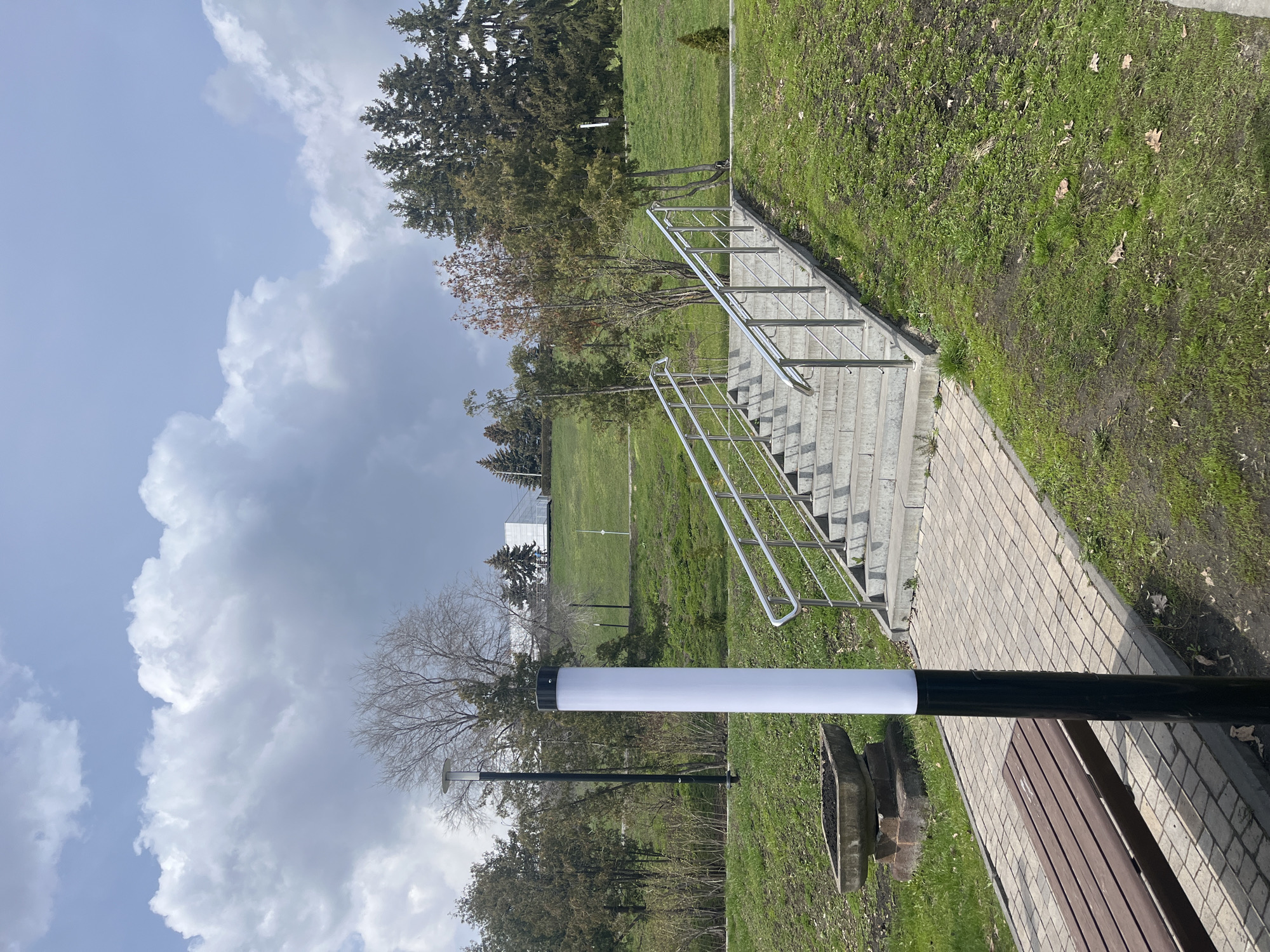 This media file depicts an outdoor park scene in Ulyanovsk, Russia, under a bright, partially cloudy sky. The image shows a paved pathway made of light-colored bricks or pavers, which runs diagonally from the lower right towards the middle. Alongside the path, a set of concrete stairs with shiny silver metal handrails ascends towards the left, leading up an embankment.
In the foreground, a modern lamp post with a black base and a tall, white cylindrical light fixture stands prominently on the left side of the path. Behind the lamp post, partially visible on the right, are a wooden slatted bench and a stone or concrete planter, suggesting a public resting area.
Extensive grassy areas flank both sides of the path and stairs, extending into the midground. The grass appears somewhat sparse in places, with visible patches of soil and scattered dry leaves, indicating a period of late winter or early spring. Various trees populate the scene, including some deciduous trees with bare branches and several evergreen conifers with dark green foliage.
In the distance, across a wide grassy field, a white building or structure is visible, along with another slender black pole, possibly a smaller lamp post. The sky above is a mix of light blue and large, cumulus clouds ranging from white to grey. The overall impression is a quiet and well-maintained public park on a bright, cloudy day. No people or visible text are present in the image.