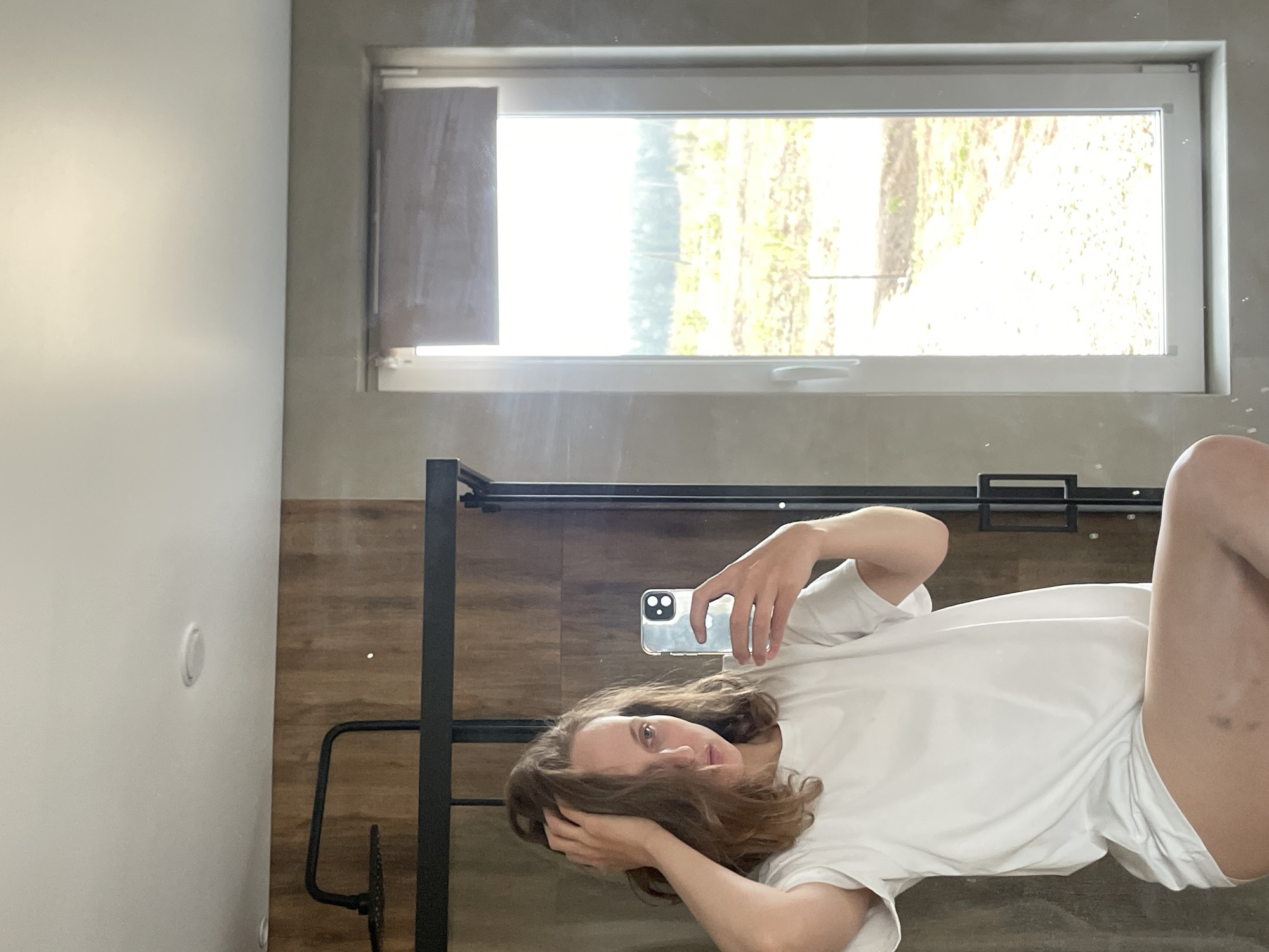The original image, oriented on its side, displays a mirror selfie of a person lying down. The individual, with long, wavy brown hair, is wearing a white t-shirt and white shorts, looking directly towards the camera with a neutral expression. They are holding a silver smartphone, likely an iPhone given the visible camera module, with a clear case in their right hand to take the photo. Dark bruises are noticeable on their upper right thigh.
The setting is an indoor room in Подшивалово, Russia, possibly a bedroom. A black metal structure, which appears to be a bed frame or furniture support, is prominent in the shot. The background features a wall divided into two sections: one is a light, off-white color with a round white fixture (potentially a light switch or smoke detector), and the other has a darker, wooden-paneled or wood-planked appearance. Above the person (when the image is correctly oriented), a rectangular window with a white frame allows bright daylight to stream in, revealing vibrant green and yellow foliage outside, suggesting a natural outdoor view during a clear daytime. The scene captures a casual, personal moment. No discernible text is visible.