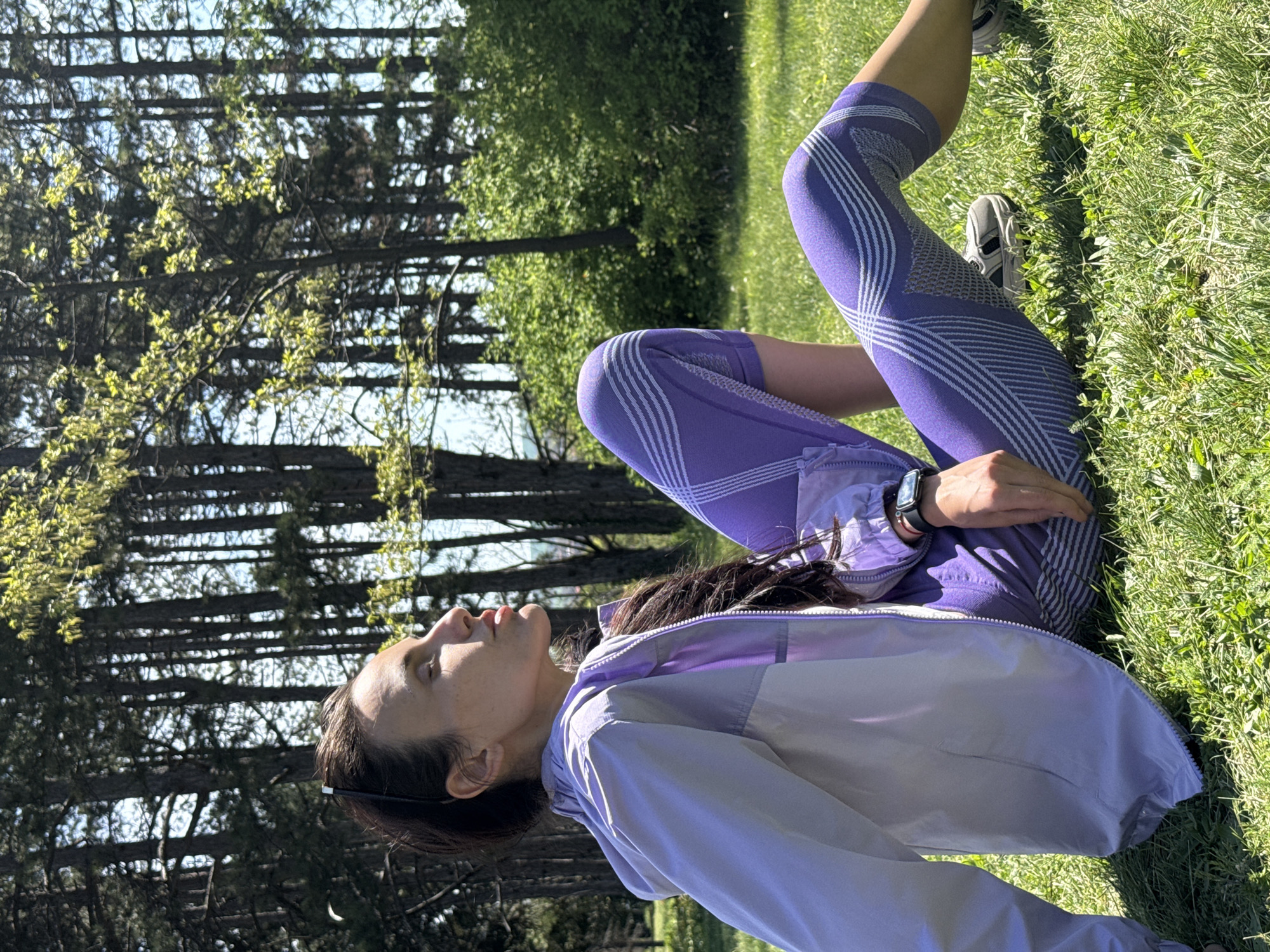 A woman is captured outdoors in Pleven, Bulgaria, seemingly resting or stretching on green grass during a sunny day. She is positioned on her back or side, with her knees bent and legs drawn up. She is wearing a light purple windbreaker jacket and matching purple athletic leggings with distinct white geometric patterns. A smartwatch is visible on her left wrist, and light-colored athletic shoes are on her feet. Her long, dark hair is loose, and her head is tilted back, with her eyes appearing to be closed or gazing upwards, conveying a sense of calm.
The setting is a natural, open area, likely a park or a field adjacent to a forested section. The foreground and midground are covered in lush green grass. In the background, a line of tall trees, including both coniferous and deciduous types, stands against a bright sky. The strong, direct sunlight creates clear shadows, indicating either morning or late afternoon. The scene evokes a peaceful and refreshing outdoor environment. No visible text is legible in the image.