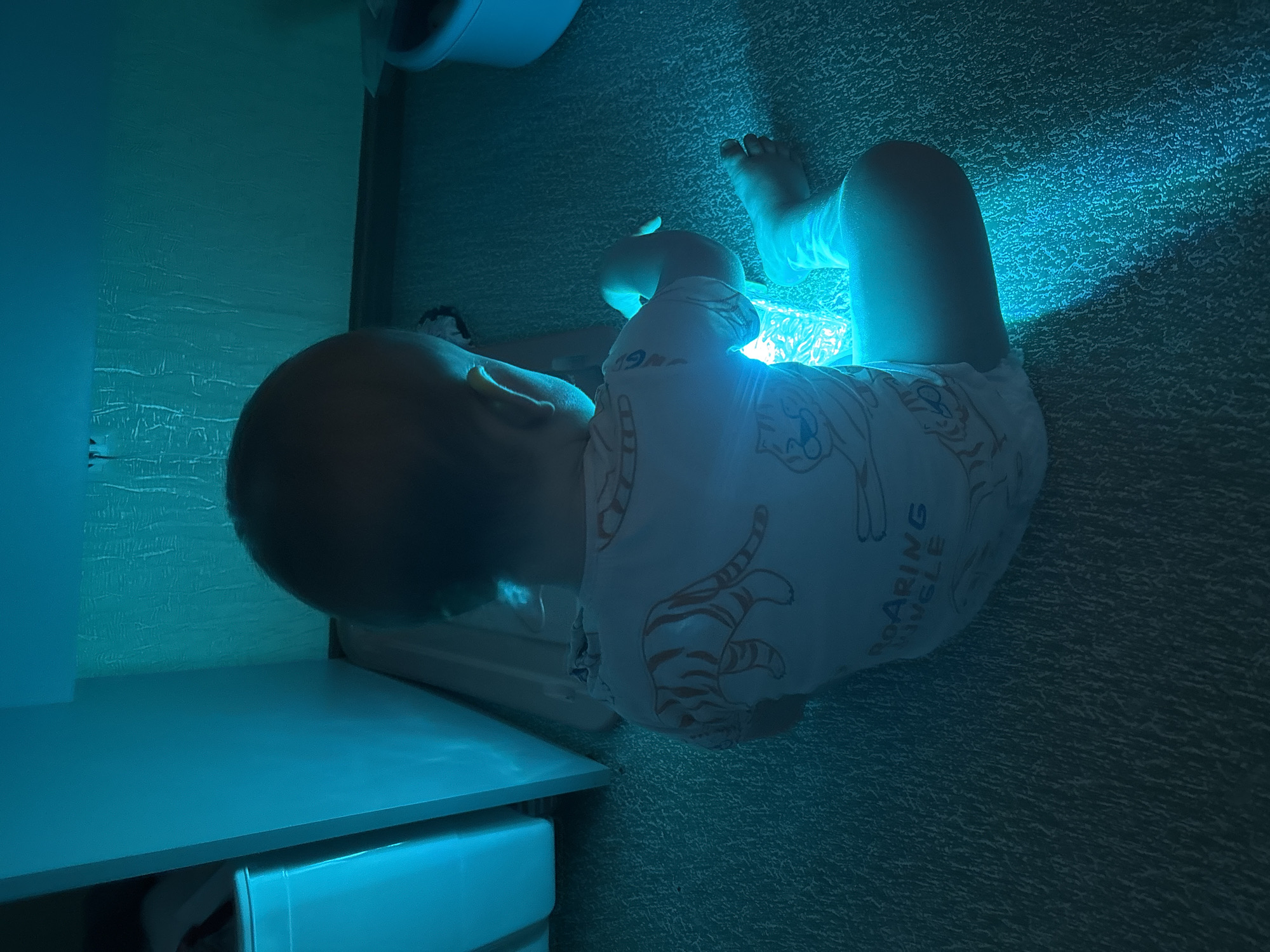 A toddler wearing a white onesie with orange tiger graphics and text that reads "ROARING JUNGLE" is sitting on a carpeted floor, illuminated by a bright blue light emanating from an object they are holding. The toddler's back is to the camera, and they appear to be looking down at the object. The environment is dimly lit, with the blue light creating strong shadows and highlighting the texture of the carpet. To the left of the toddler, a white cabinet or shelf is visible, also bathed in blue light. The scene suggests an indoor play or relaxation setting. The provided image does not contain enough visual information to determine its specific location within Naberezhnye Chelny, Russia.