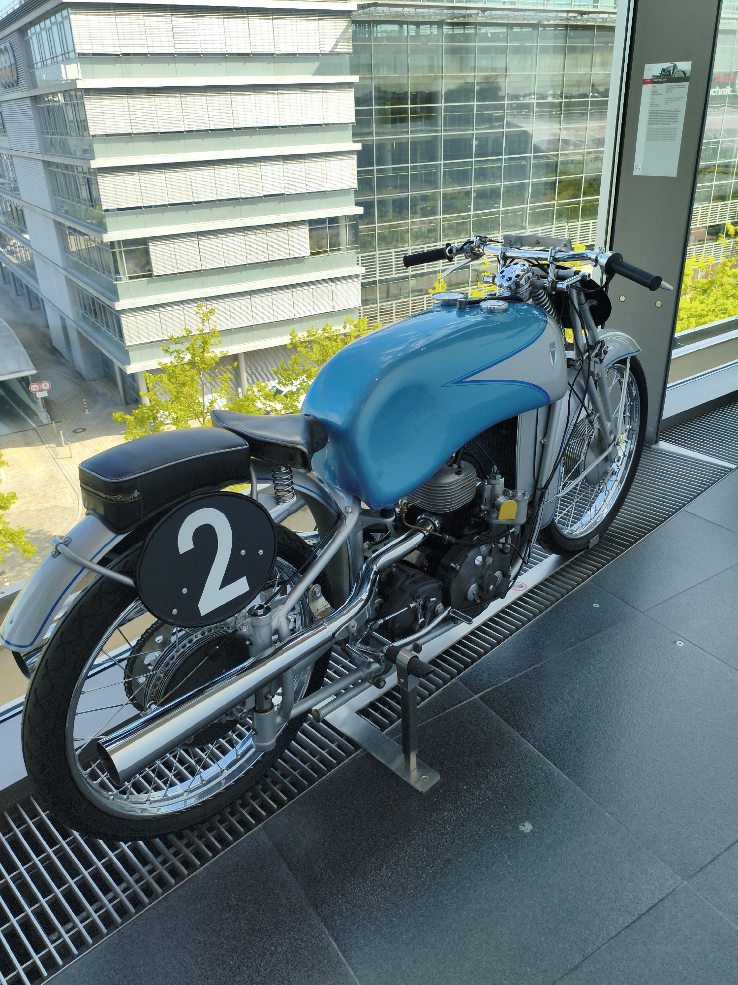 This image displays a vintage blue and silver motorcycle with the number "2" on a circular black plate positioned on its rear fender. The motorcycle is situated indoors, likely in a museum or exhibition space. Behind the motorcycle, a modern glass-fronted building with horizontal blinds is visible, reflecting the surrounding greenery and sky. The flooring consists of dark grey tiles and a metal grate. A display panel with text and an image is mounted vertically to the right of the motorcycle. Based on the context of the building's architecture and the presence of the motorcycle which is a BMW R50, this scene is likely located at the BMW Museum in Ingolstadt, Germany. There are no people visible in the image.