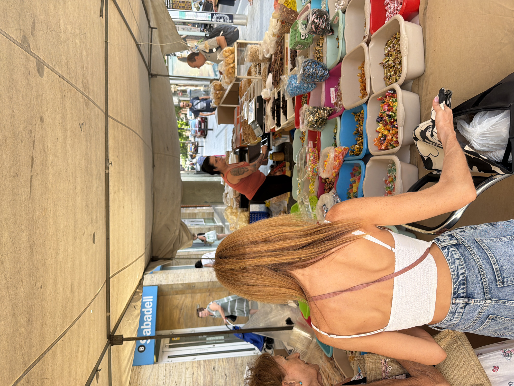 A street market stall is set up under a beige tarp supported by metal poles. The stall displays a variety of candies and dried fruits in numerous plastic bins. In the foreground, a woman with long, reddish-brown hair is viewed from behind, wearing a white crop top and denim shorts. She is reaching into a black tote bag. To her left, another woman with short brown hair and a turquoise earring is partially visible, holding a straw bag. Behind them, a woman in an orange top is attending to the stall, and a man in a grey shirt is interacting with her, holding a bag of snacks. In the background, a street is visible with buildings, cars, and pedestrians. A sign for "Sabadell" is mounted on a building to the left. The overall scene depicts a bustling outdoor market.