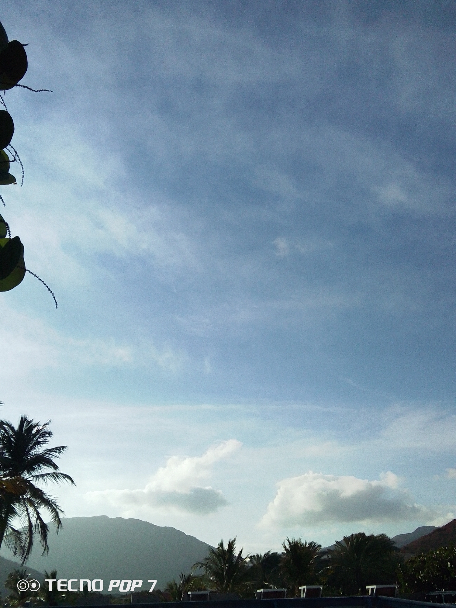 The image shows a view of the sky and landscape in Pedro González, Venezuela. The sky is mostly blue with wispy clouds. In the lower portion of the image, there are mountains, trees, and palm trees. A building with a wall is visible near the bottom of the image. The camera phone used for the shot is a TECNO POP 7.