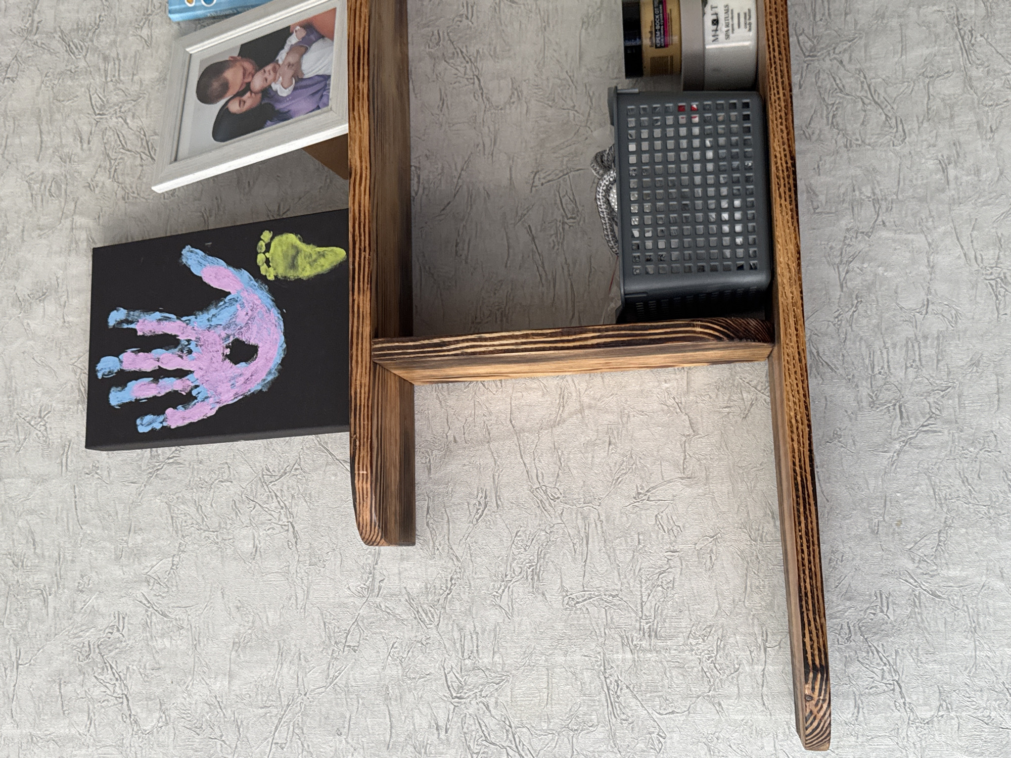 A framed family photo and a child's handprint artwork are displayed on a shelf against a textured light gray wall. The photo shows a man, a woman, and a baby, with the man kissing the baby. The handprint artwork is on a black canvas and features purple and blue paint. A yellow footprint is also visible on the canvas, above the handprint. To the right, on the same shelf, are various cosmetic products, including jars and bottles with visible text that includes "SPA RITUALS". A gray, lidded container with a grid pattern is also on the shelf, connected to a thin rope or cord. The shelf itself appears to be made of wood with a burnt or stained finish. The overall scene suggests a home environment, possibly a bathroom or dressing area, where personal mementos and toiletries are kept. The lighting is neutral, suggesting indoor illumination.