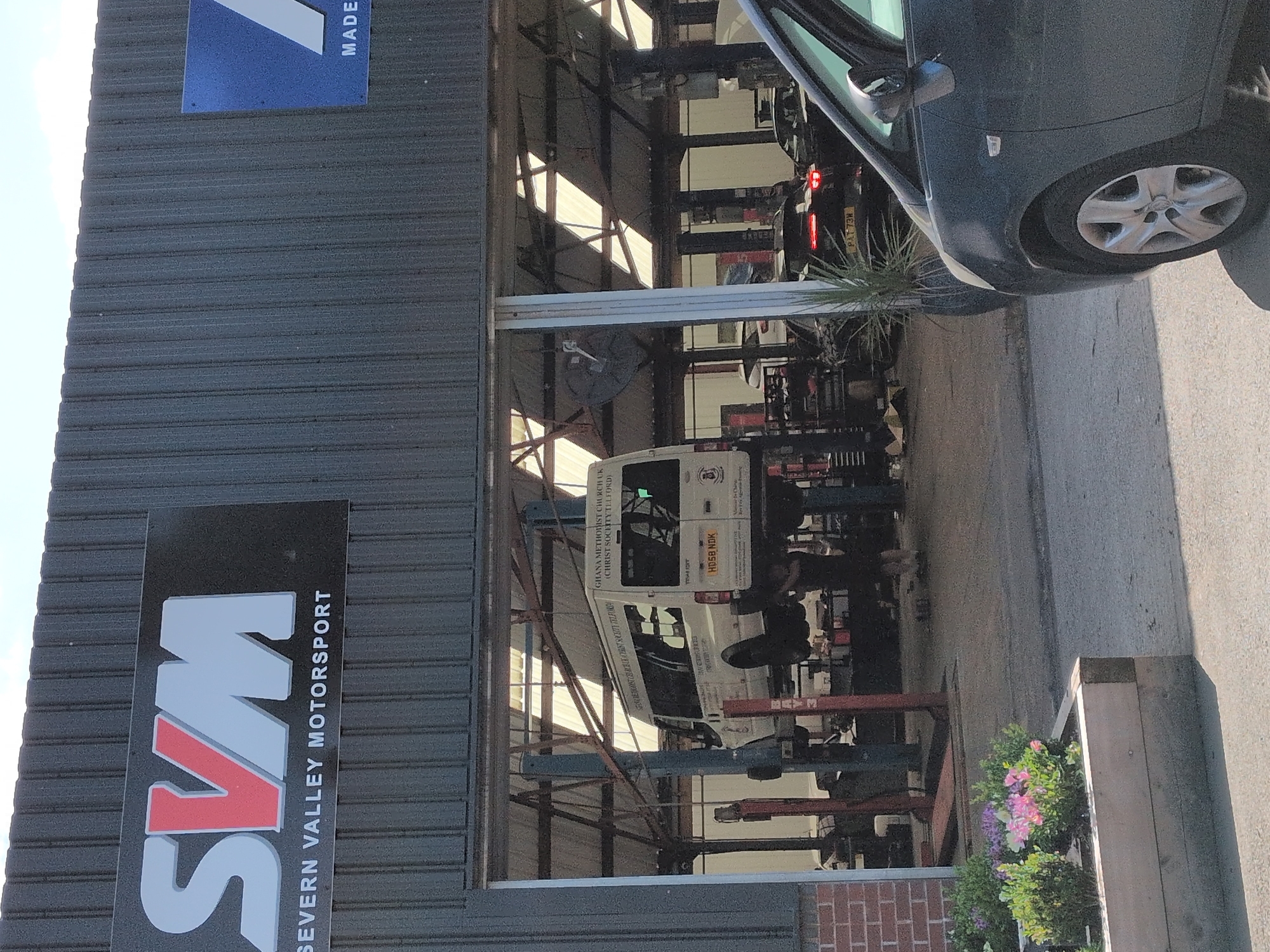 This is a horizontal, eye-level, outdoor shot of a motor vehicle repair shop. The shop appears to be a garage with a metal exterior and a corrugated metal roof. Inside the garage, several vehicles are on car lifts. A dark grey car is parked outside the garage to the right, and a white van with text on its side is visible on a lift inside. The van has text that reads "GHANA METHODIST CHURCH UK (CHRIST SOCIETY TLLFORD)". The license plate on the van is "HDSB NDK". There is a sign on the exterior of the building that says "SVM SEVERN VALLEY MOTORSPORT". Another sign in the upper left corner has the word "MADE" on it. The scene is well-lit, suggesting it's daytime, possibly sunny. There are some plants in a planter box to the right of the garage. The overall impression is a busy, functional repair shop.
