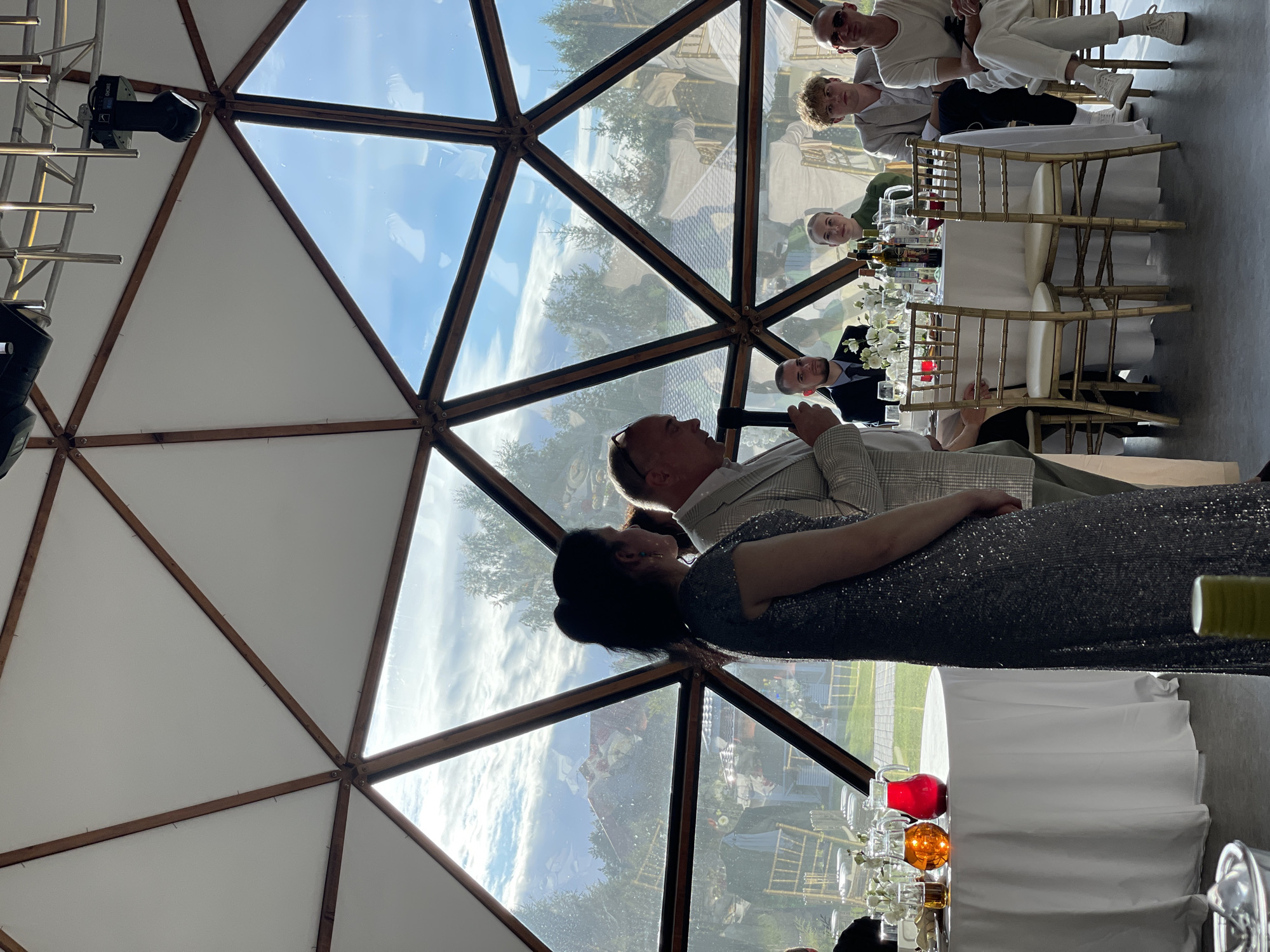 A man in a light-colored plaid suit is speaking into a microphone, standing next to a woman in a sparkling dark dress. They are inside a geodesic dome structure with large triangular glass panels that offer a view of trees and buildings outside under a partly cloudy sky. Several people are seated at a round table in the background, some with drinks and bottles on it. A white tablecloth covers the table. The floor appears to be concrete. The scene suggests an indoor event or reception.