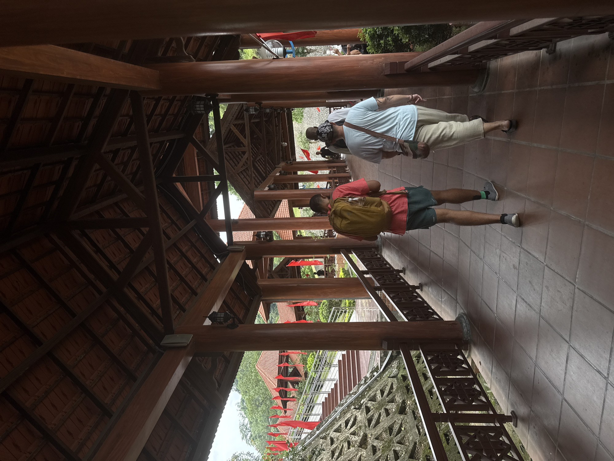 A covered walkway in Đồng Đăng, Vietnam, features a tiled floor and wooden columns supporting a red-tiled roof. Two individuals are walking along the walkway. One wears a pink shirt, gray shorts, and carries a yellow backpack. The other wears a blue shirt and tan pants. The walkway's side is lined with a decorative iron railing overlooking stairs. Red flags are visible outside. The overall construction style is traditional Vietnamese architecture.
