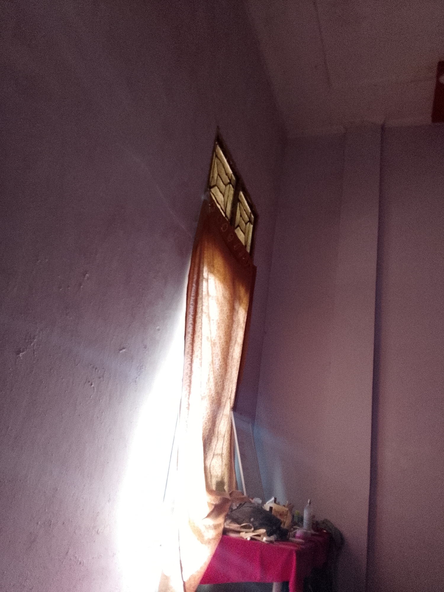 The image depicts an indoor scene within a building located in Lhokseumawe, Indonesia. No individuals are present. The room features purplish-grey walls, one of which exhibits textural inconsistencies and minor imperfections on the left side. A multi-paned window with a distinct geometric pattern is positioned high on the wall, partially obscured by a light brown or beige curtain. A powerful, concentrated beam of natural sunlight enters from the left, casting a bright diagonal highlight across the left wall and intensely illuminating the curtain. To the lower right, beneath the window area and partially obscured by the curtain, a small table draped with a bright red fabric is visible. This table holds an assortment of objects, including a white bottle or container, various fabric items or clothing, and other unidentifiable miscellaneous articles, suggesting a modest and functional arrangement. The right side of the room, including a corner and a vertical architectural feature, remains in comparative shadow.