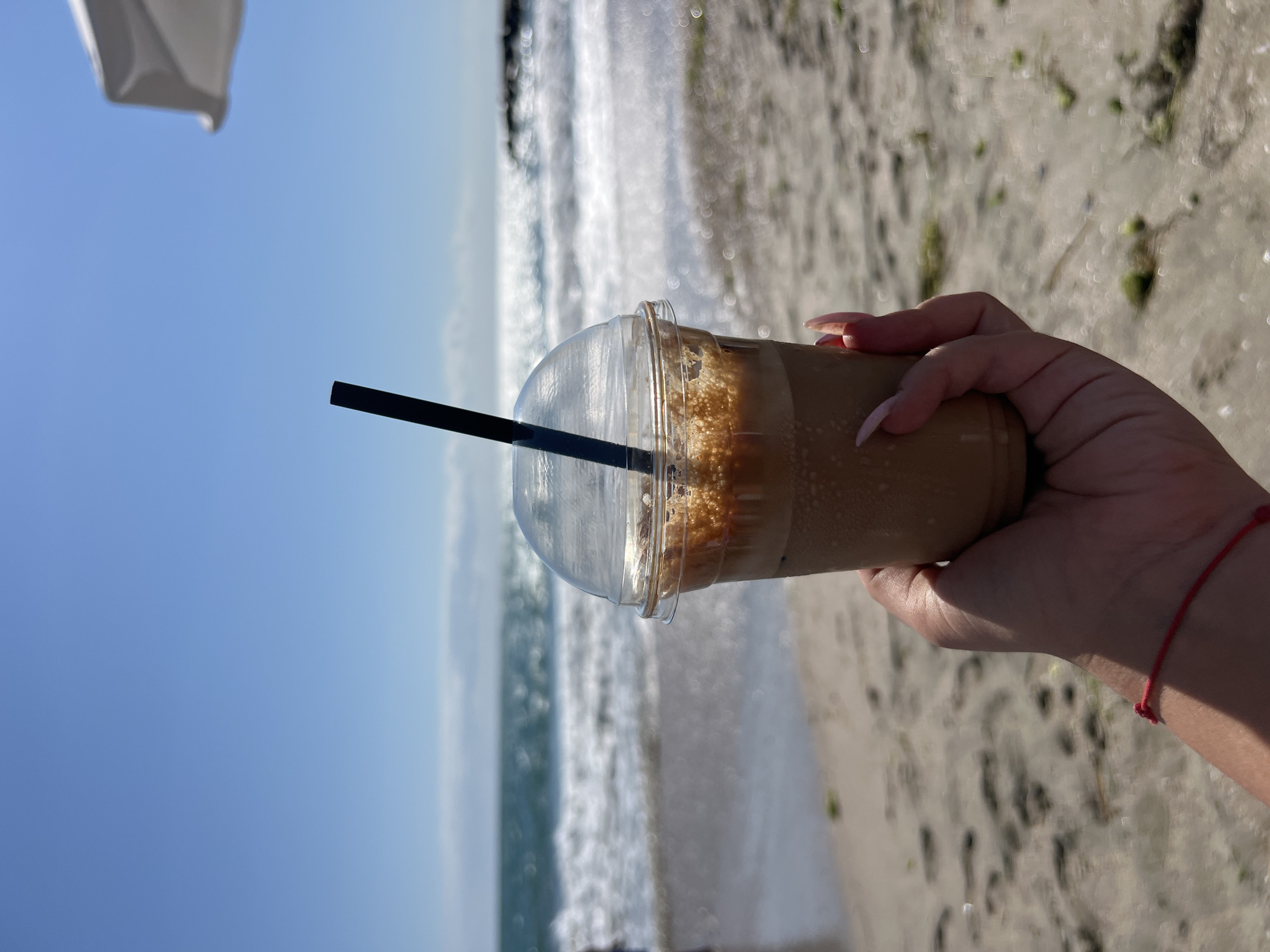 A hand with long, light-colored fingernails holds a clear plastic cup filled with a brown, frothy beverage. A black straw extends from the cup. The cup is positioned in the foreground, against a blurred background of a beach and the sea. The sand appears light brown with scattered patches of green. Gentle waves are visible in the turquoise sea, and the sky is a clear blue with some hazy white clouds near the horizon. A hint of a white umbrella is visible in the upper right corner. This scene is likely the beach in Pomorie, Bulgaria, given the typical landscape and beach umbrella common in tourist areas.
