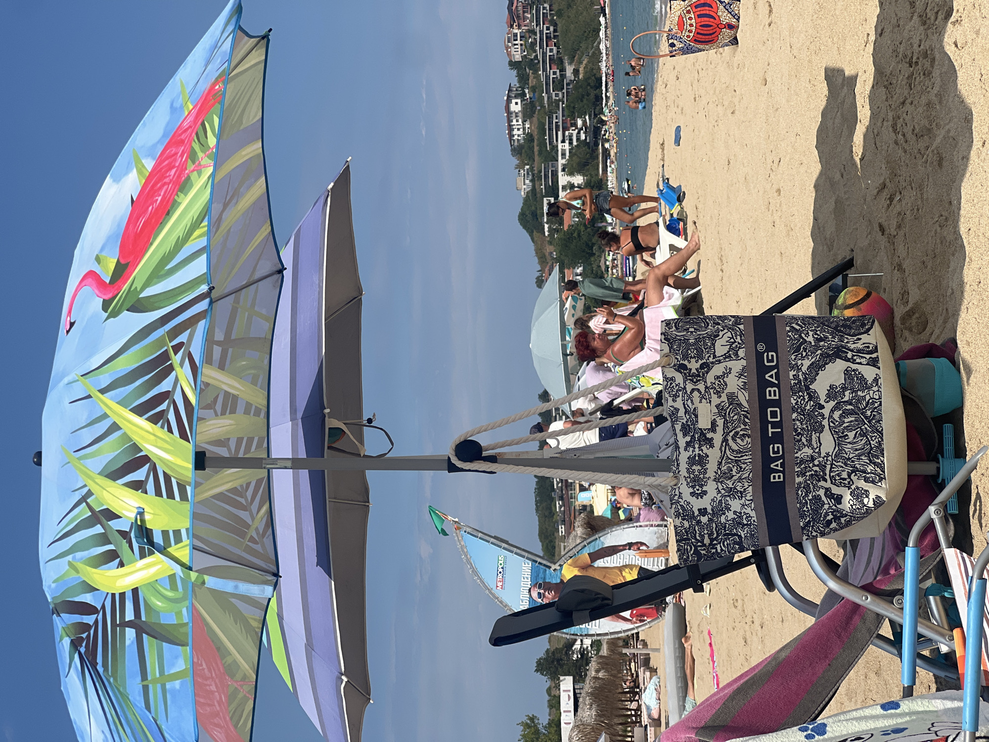 This image captures a sunny day on a beach in Sozopol, Bulgaria. In the foreground, a beach chair is partially visible, draped with a striped towel. A large tote bag with a blue and white pattern featuring the text "BAG TO BAG" is hanging from a beach umbrella pole. The umbrella itself has a vibrant tropical design with flamingos and palm leaves.
Further back on the sandy beach, several people are relaxing. Some are seated on beach chairs, while others are standing. In the middle ground, a man in a yellow shirt and sunglasses stands near a large sign for "METROPOLIS OUTDOOR MARINE GROUP". Beyond the beach, the calm sea is visible, dotted with numerous people swimming and enjoying the water. In the background, houses are built on a hillside overlooking the beach and the sea, indicating the urban setting of Sozopol. The overall atmosphere is one of leisure and a typical summer day at the beach.