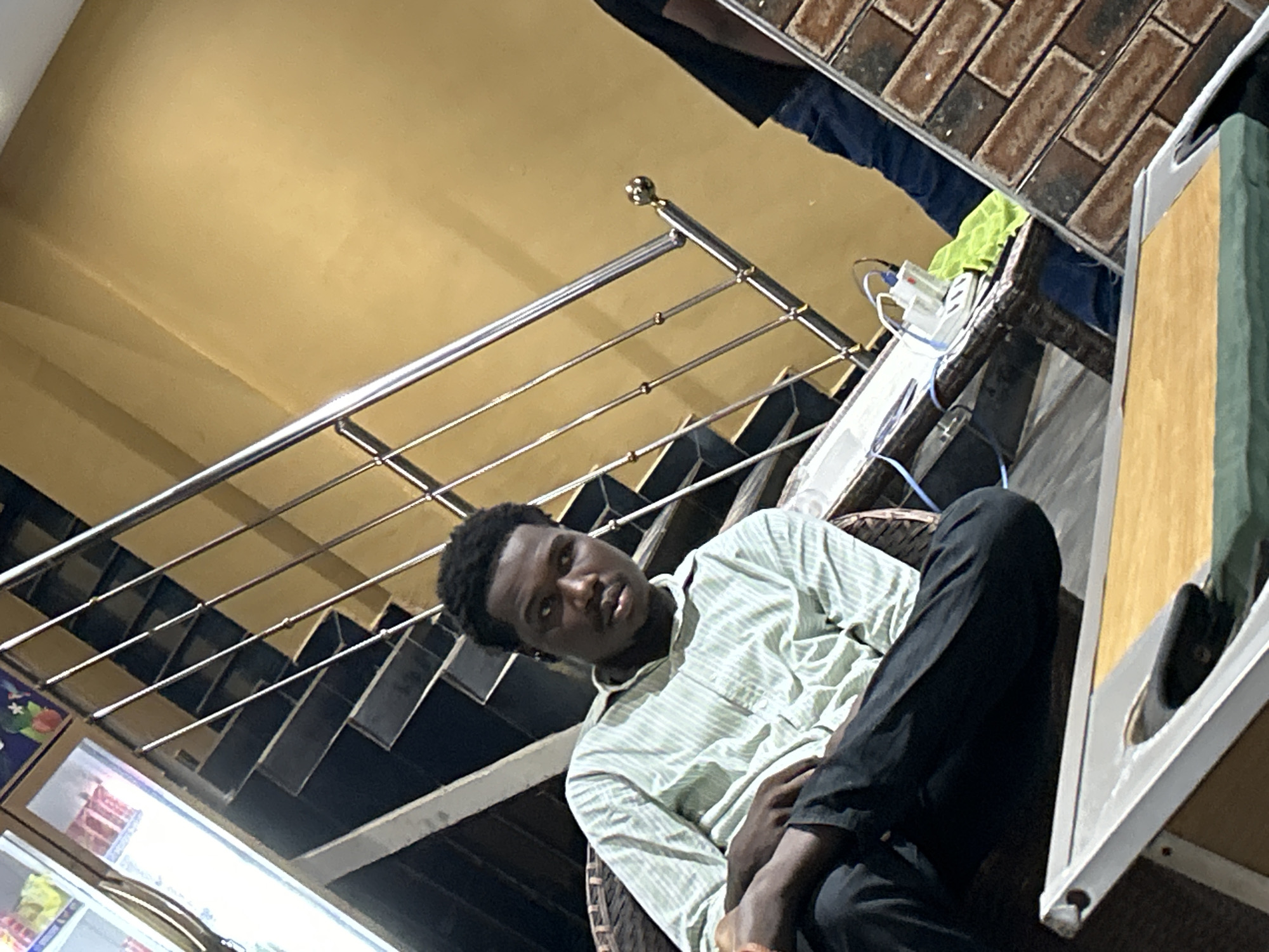 A young Black male is seated indoors, appearing relaxed, with his left arm resting on his lap. He is wearing a light-colored, striped button-down shirt and dark trousers. The background features a modern staircase with a metal railing and a yellow-painted wall. To the right of the staircase, a brick-patterned wall is visible, and a table with electrical outlets and a bright green cloth is present. The scene suggests an indoor setting, possibly a residential or commercial space in Ihiagwa, Nigeria.