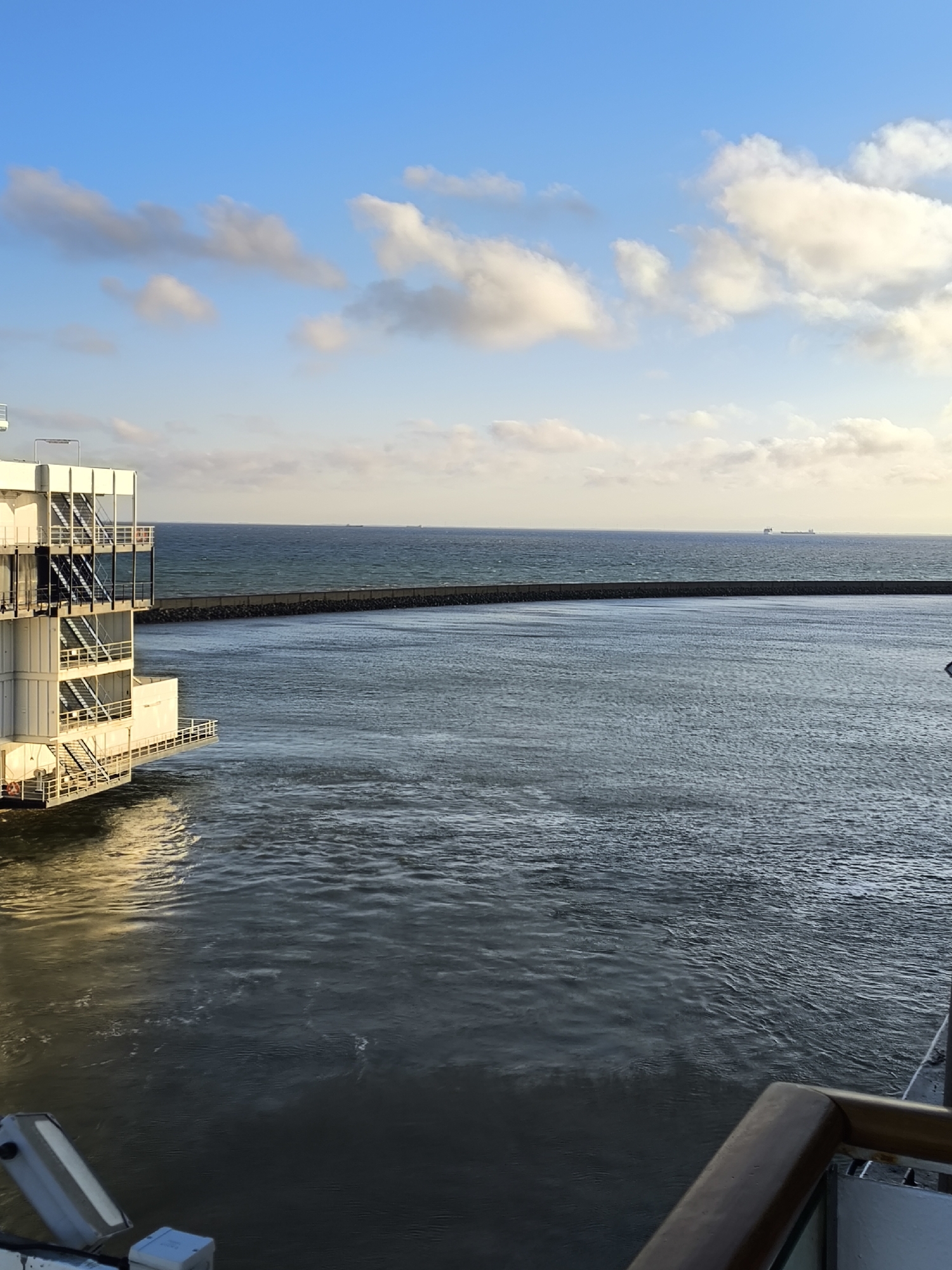 The image depicts a view of the ocean from a raised vantage point, possibly a cruise ship or a pier, in Fehmarn, Germany. On the left side, a modern, white, multi-story structure with visible staircases and balconies extends into the water. This structure appears to be an industrial or maritime facility. The water is choppy and dark, with sunlight reflecting off its surface. In the distance, a long, dark breakwater stretches across the horizon, separating the inner harbor from the open sea. Beyond the breakwater, several cargo ships are visible on the vast expanse of the ocean. The sky is a clear blue, dotted with fluffy white clouds, suggesting fair weather. The overall scene conveys a sense of maritime activity and coastal landscape.