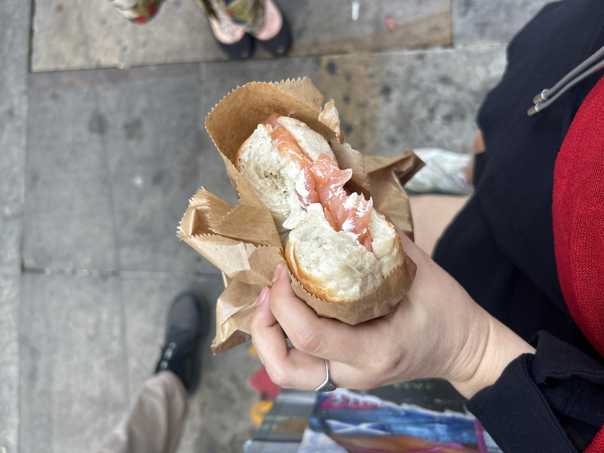 A person's hand, adorned with a silver ring, holds a salmon and cream cheese bagel wrapped in brown paper. The bagel appears to be sliced in half and filled generously with pink salmon and white cream cheese. In the background, blurred figures are visible, including a person wearing black shoes and another person wearing patterned shoes. The ground is paved with grey rectangular stones. The overall scene suggests an outdoor setting, possibly a street or public square in London, United Kingdom.