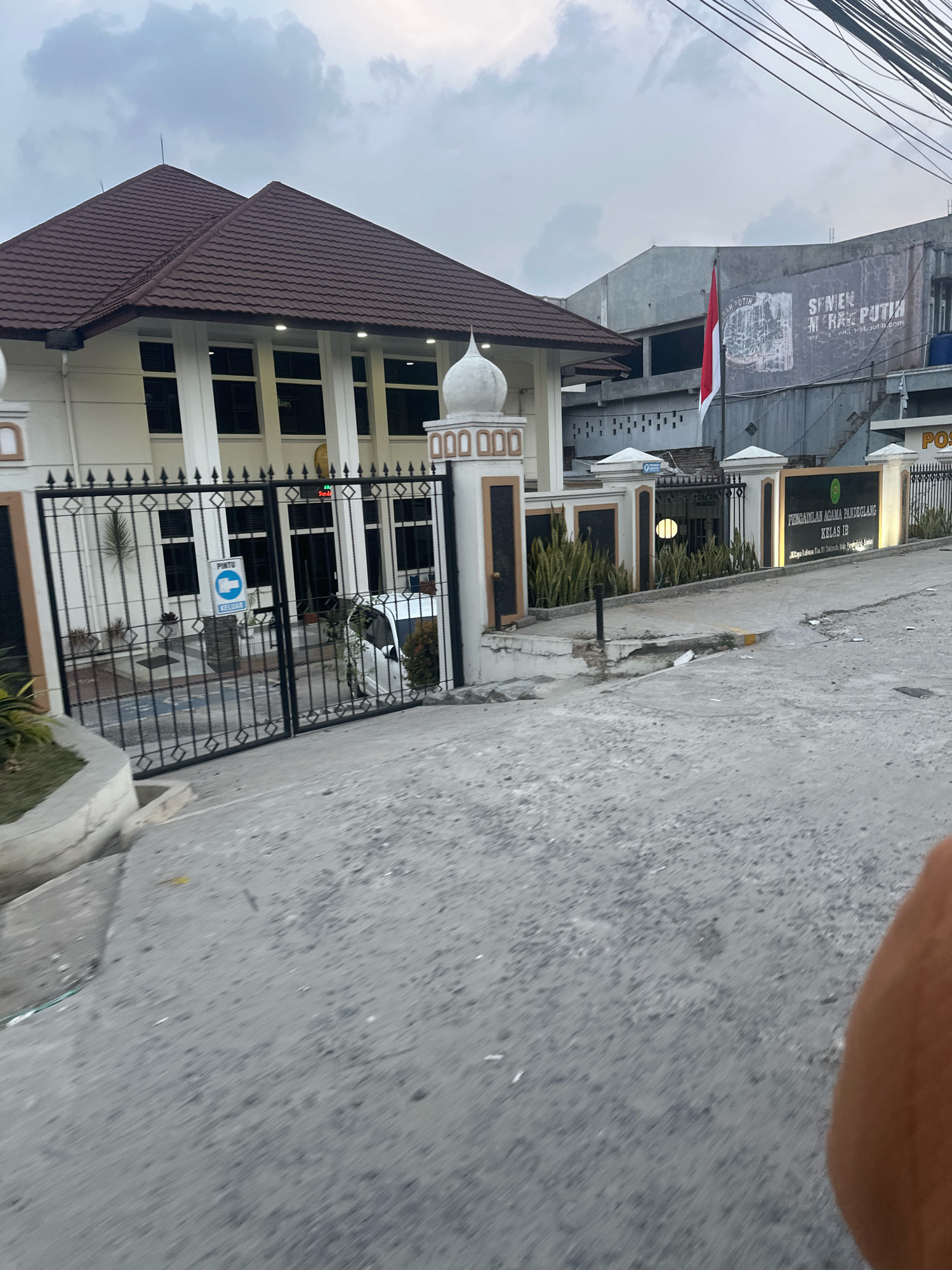 The image shows a wide shot of the *Pengadilan Agama Pandeglang Kelas IB* in Saruni, Indonesia. The building is white with a brown tiled roof and tall windows. A black metal gate with decorative elements is in front of the building. A sign with the building's name and class is visible on a white pillar. The street in front of the building is paved with concrete. A white car is parked inside the gate. A red and white Indonesian flag is flying from a pole on a building in the background. The sky is overcast. There are no people visible in the image.