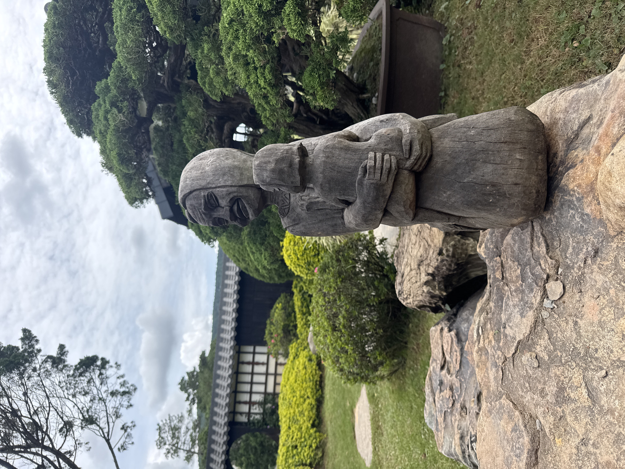 A wooden sculpture of two figures, possibly a mother and child, sits on large rocks in an outdoor garden setting. The figures are carved from dark wood and appear to be embracing. The sculpture is weathered, showing cracks and the grain of the wood.
The background features lush greenery, including manicured bushes and a large, gnarled tree. In the distance, a traditional-looking building with a dark roof and latticed windows is visible. The setting suggests a garden or park, possibly in a temperate climate given the foliage.
The sky is overcast with clouds, indicating a cloudy day. The lighting is soft, without harsh shadows. The overall mood is serene and contemplative, perhaps evoking a sense of nurture or companionship. There are no visible signs of text in the image. The location context is Cam Ly, Đà Lạt, Vietnam.