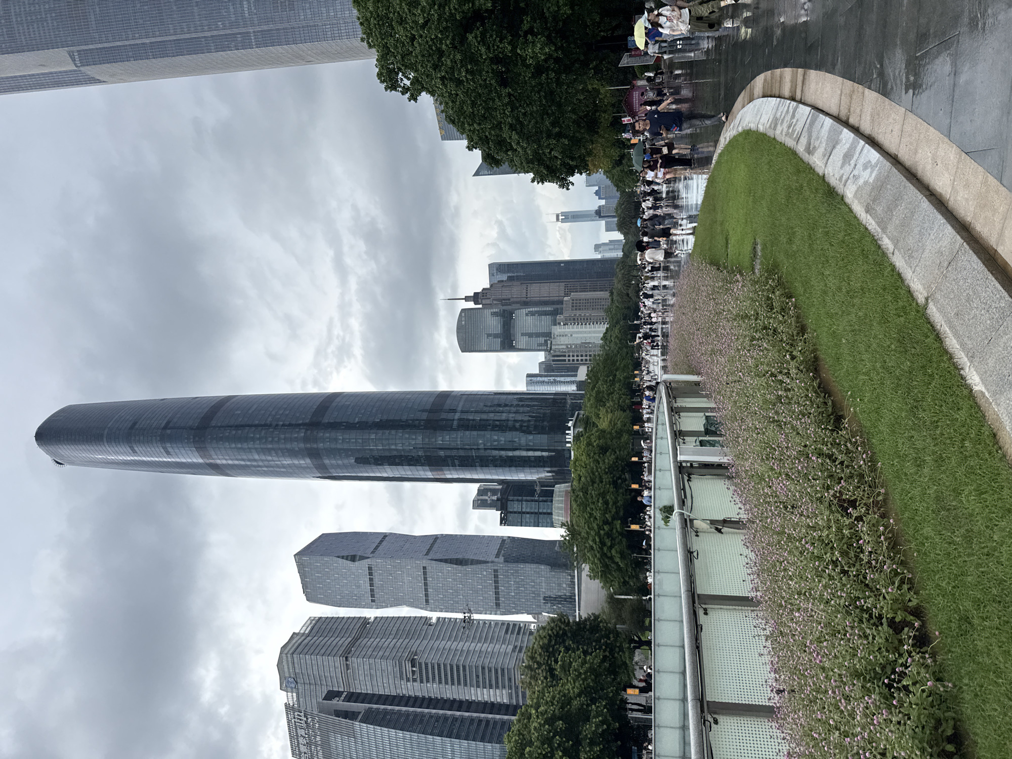 The image depicts a cityscape in Tianhe District, China, characterized by a dense collection of modern skyscrapers under an overcast sky. A wide, wet pedestrian walkway occupies the foreground and middle ground, with a curved grassy area and a flower bed to the left. Numerous people are walking along the wet path, some carrying umbrellas, indicating recent or ongoing rain. Several tall, sleek buildings with glass facades dominate the skyline, with a particularly prominent, slender skyscraper in the center. Trees line the walkway and separate the pedestrian area from the buildings. The wet pavement reflects the light from the overcast sky and surrounding structures.