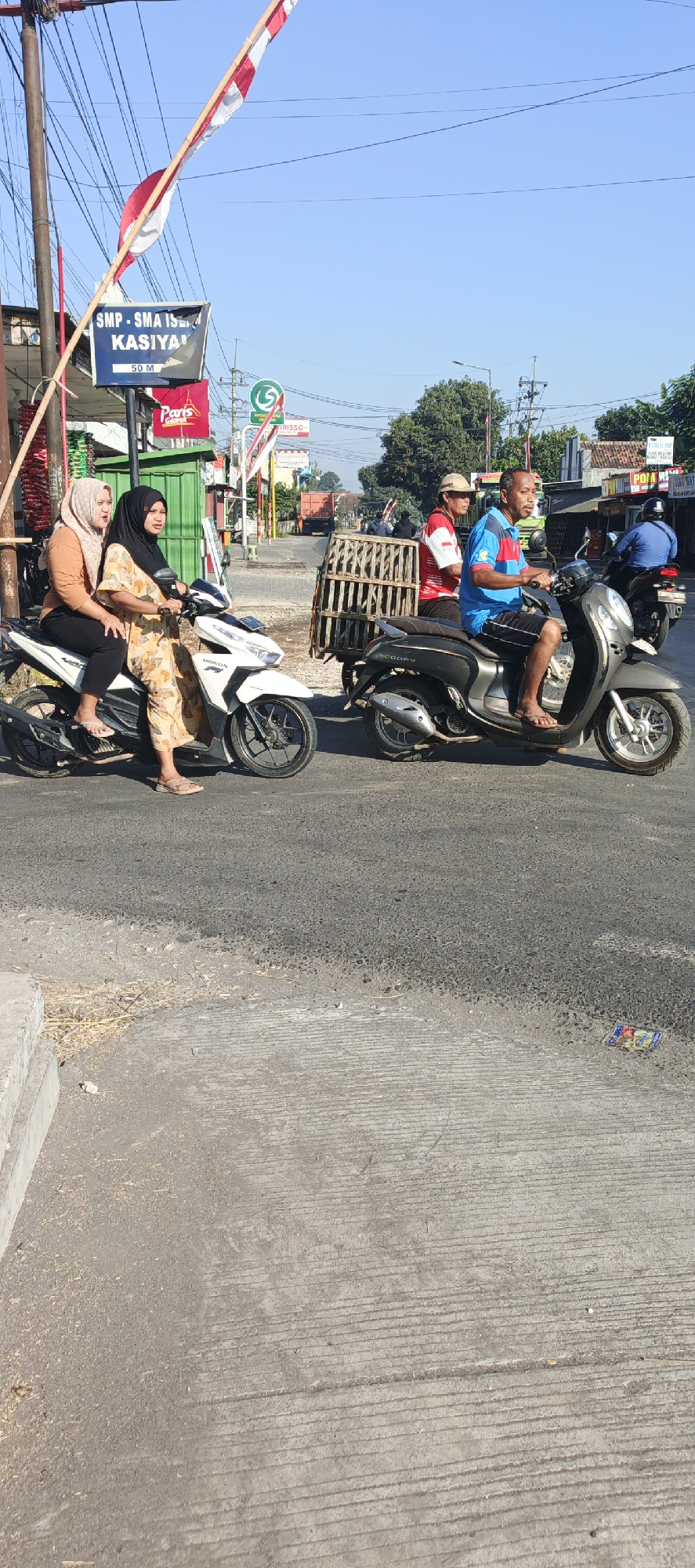 The image depicts a street scene in Kasiyan, Indonesia, on a bright, clear day. In the foreground, two women are riding a white scooter. The woman in the back is wearing a light brown hijab and an orange shirt, while the woman in the front is wearing a black hijab and a patterned yellow dress. They are both looking forward. To the right, a man wearing a blue and red striped shirt and black shorts is riding a dark gray scooter. He is looking ahead. Behind him, another person on a scooter is partially visible. A wooden crate is attached to the back of the dark gray scooter. In the background, there are buildings, trees, utility poles with wires, and a blue sky. A sign for "SMP - SMA ISLAM KASIYAN" with "50 M" is visible on the left, indicating the location. Other smaller signs and some construction or fencing are also present in the background. The road surface is asphalt, with a concrete sidewalk or curb on the left.