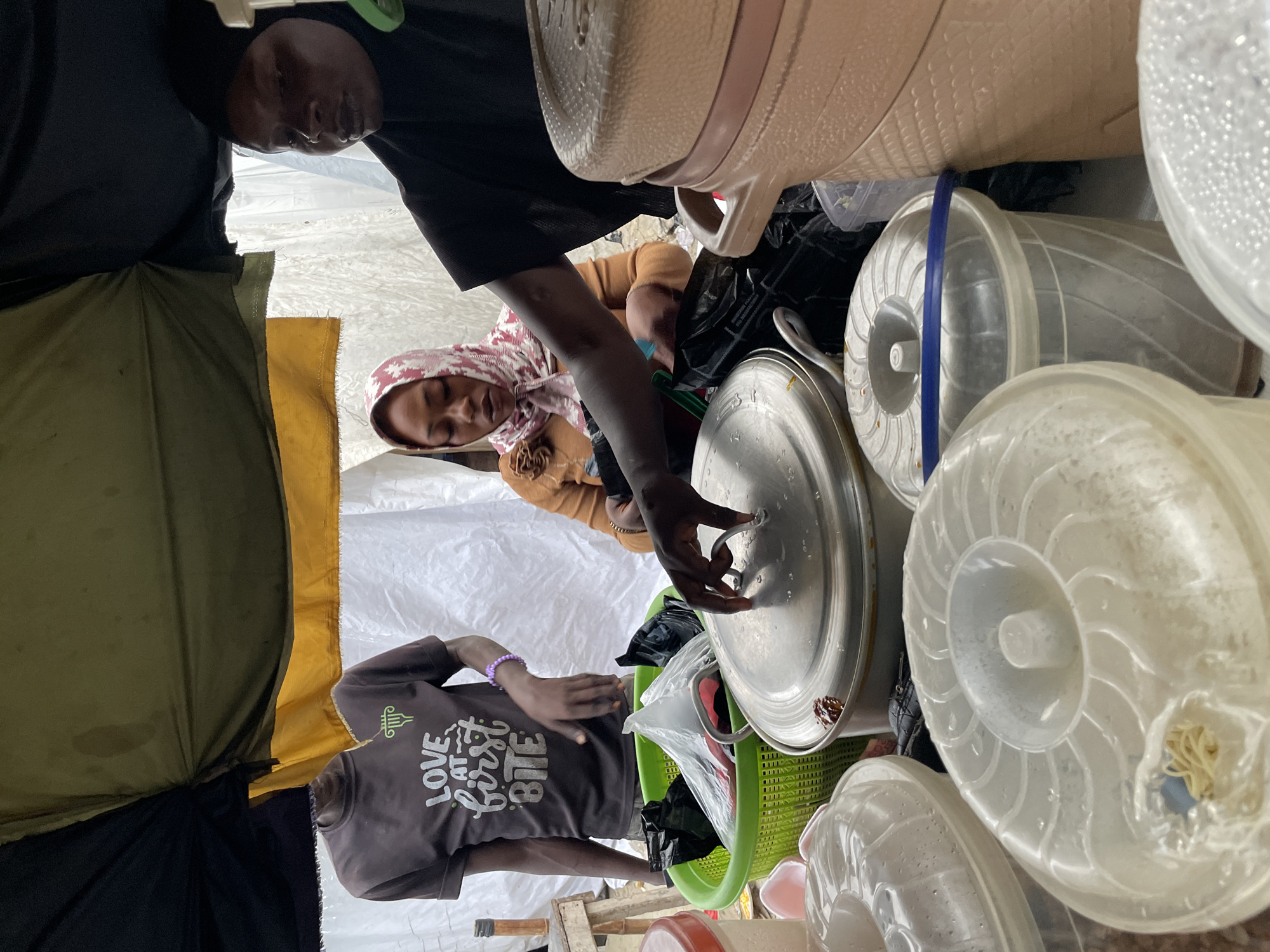 In Ilado, Nigeria, under a makeshift canopy of green and yellow fabric, a bustling scene unfolds. A young person wearing a dark t-shirt with the words "LOVE AT FIRST BITE" stands near a bright green plastic basket filled with black plastic bags. To their right, a woman in a tan top and a patterned headscarf is focused on something in her hands. Further to the right, another woman, wearing a black hijab, is partially visible, her attention directed towards a large, beige, cylindrical container stacked with several smaller containers. In the foreground, several transparent plastic containers with white lids are arranged, one showing a small amount of spaghetti-like food. A large, metallic pot with a lid is positioned centrally, with a hand reaching towards it, suggesting ongoing food preparation or service. The environment appears to be an outdoor market or a food stall, with a white tarp serving as a backdrop.