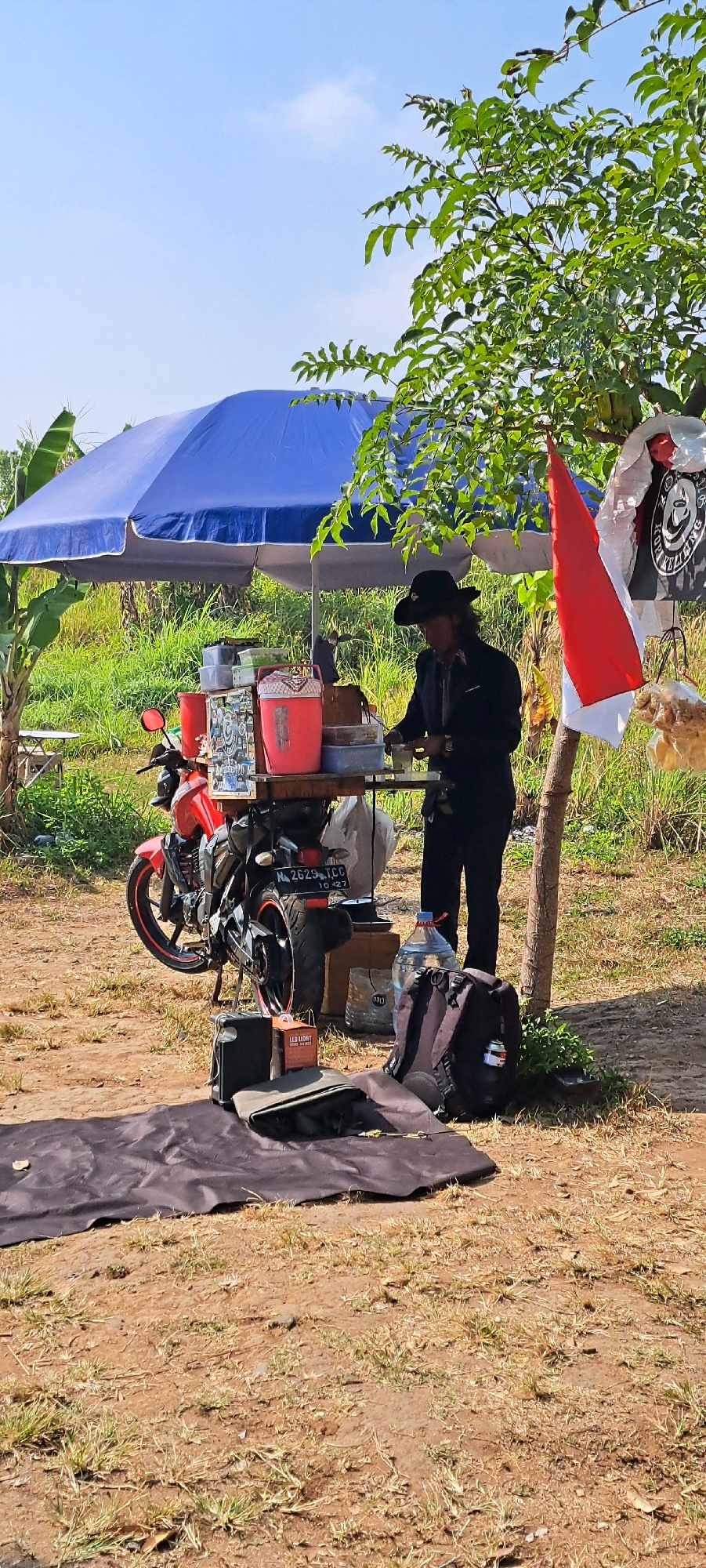 In Sekarjoho, Indonesia, on a sunny day, a person in a black cowboy hat and black clothing is operating a street food cart attached to a red motorcycle. The cart is situated under a large blue umbrella, with a red and white Indonesian flag hanging from a nearby tree. The scene is set in a rural or semi-rural environment, with tall green grass and trees surrounding the area. In the foreground, there is a dark mat with some items laid on it, including a black bag and a small box labeled "LED LIGHT". Various containers and supplies are visible on the cart, including red buckets and plastic tubs. A large bottle of water stands next to a backpack. The ground is made of dirt with patches of dry grass.