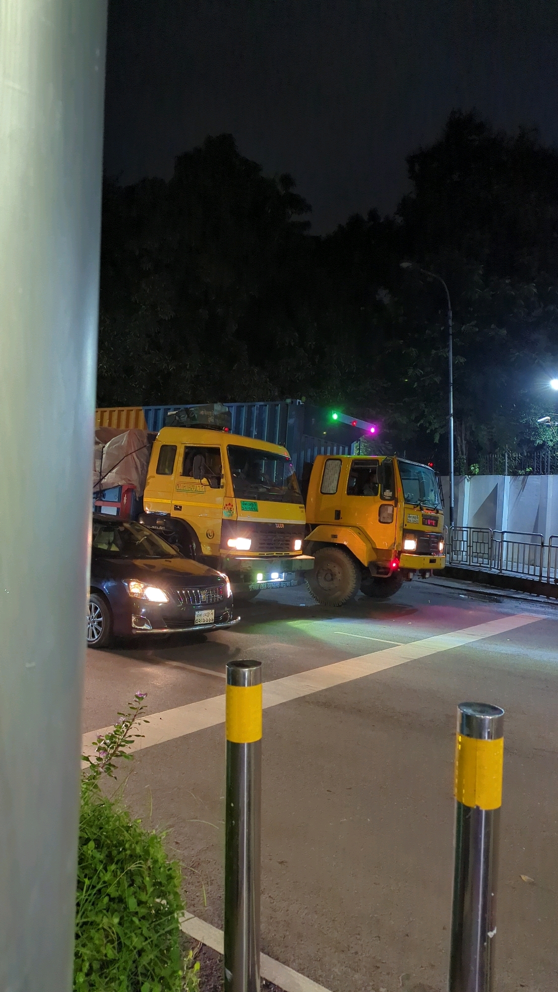 In this nighttime photograph, two yellow trucks are parked on a road in Dhaka, Bangladesh, illuminated by streetlights and the headlights of a dark-colored sedan in the foreground. The truck on the left appears to be a container truck, loaded with goods covered by a tarp. The truck on the right is a smaller flatbed truck. Both trucks have their headlights on. The sedan, facing left, has its headlights and taillights illuminated. The scene takes place at an intersection, indicated by the presence of traffic lights and painted road markings. A metal fence and lush green bushes line the right side of the road. Two metal bollards with yellow reflective bands are positioned on the road. The sky is dark, suggesting it is late at night. The overall ambiance is that of a busy urban environment at night.