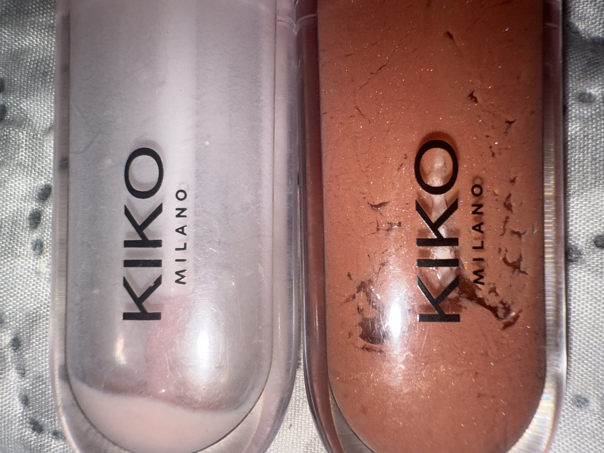 This close-up image displays two transparent tubes of KIKO MILANO cosmetic products, likely lip glosses or liquid makeup, resting on a textured, light grey fabric surface. The scene is static, focusing entirely on the products.
On the left, one tube contains a pale, pearlescent liquid with a hint of pink or white, giving it an iridescent sheen. On the right, the second tube holds a reddish-brown, glittery liquid. The glitter particles are distinctly visible within the product. Both tubes clearly show the brand name "KIKO" in large, black, sans-serif letters, with "MILANO" written vertically beneath it in a smaller font.
Notable details include the clear plastic of the tubes, allowing the texture and color of the products to be seen. The reddish-brown product appears to have some dried residue or streaks on the inside of the tube, suggesting prior use. The background fabric has a woven pattern with small, darker specks, possibly part of a blanket or soft surface. The lighting is soft, highlighting the reflective qualities of the products. While the image itself offers no visual cues to its location, the provided context indicates it was taken in Varna, Bulgaria.