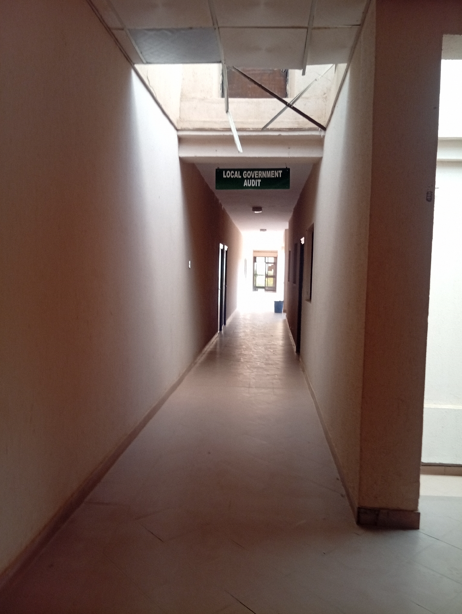 The image shows a long, narrow indoor hallway with neutral-colored walls. A sign hanging from the ceiling reads "LOCAL GOVERNMENT AUDIT." The hallway is illuminated by overhead lights and natural light coming from the far end, which opens to a brighter area with a window and a blue bin. There are doors visible on both sides of the hallway. The floor is tiled. The scene appears to be within a government building in Dutse, Nigeria.