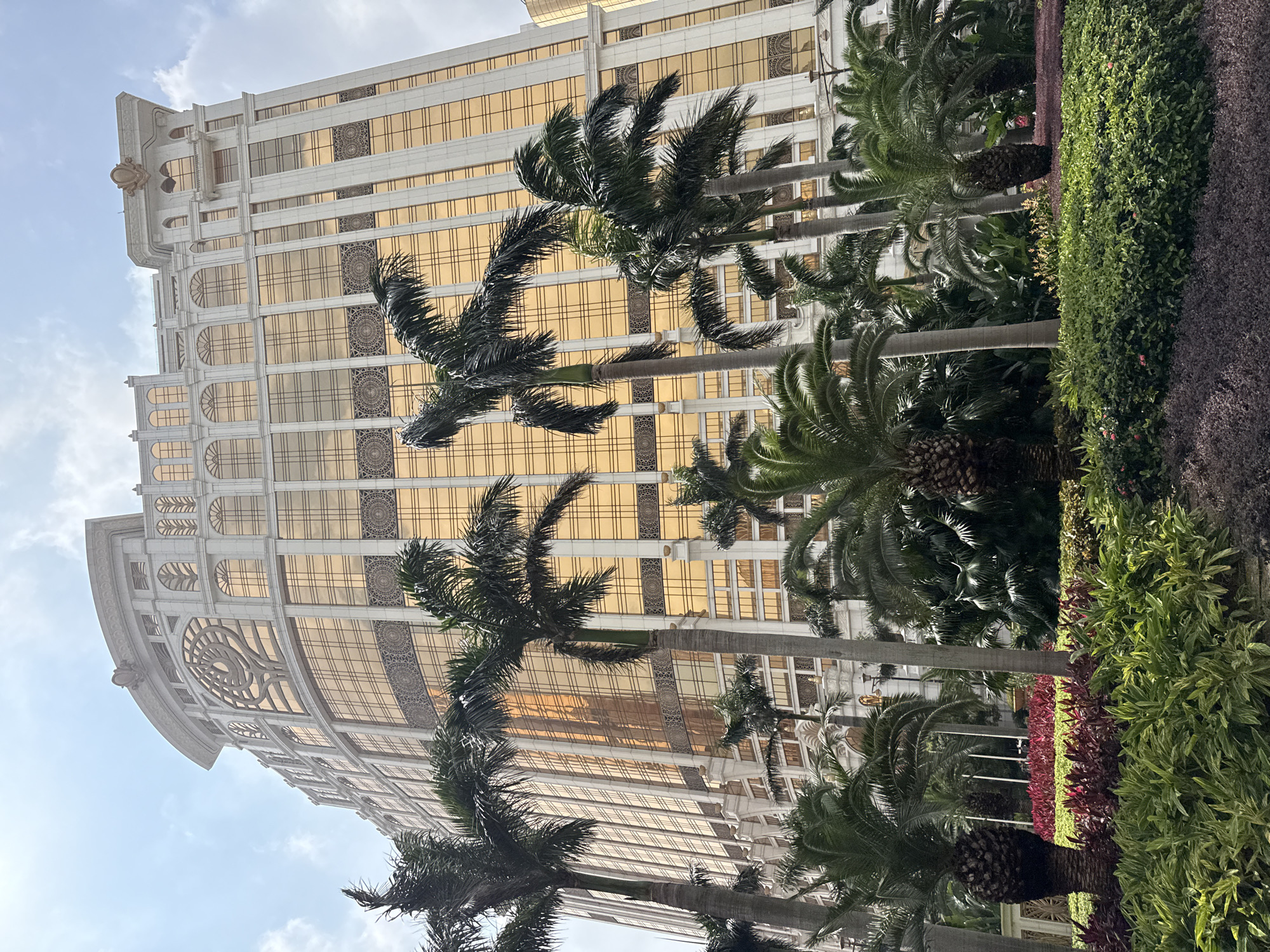 A low-angle shot depicts the Grand Lisboa Palace hotel in Macau. The hotel, predominantly gold and white, is visible through a series of palm trees and lush greenery. The foreground features a well-maintained garden with varied plant life. The sky is partly cloudy.