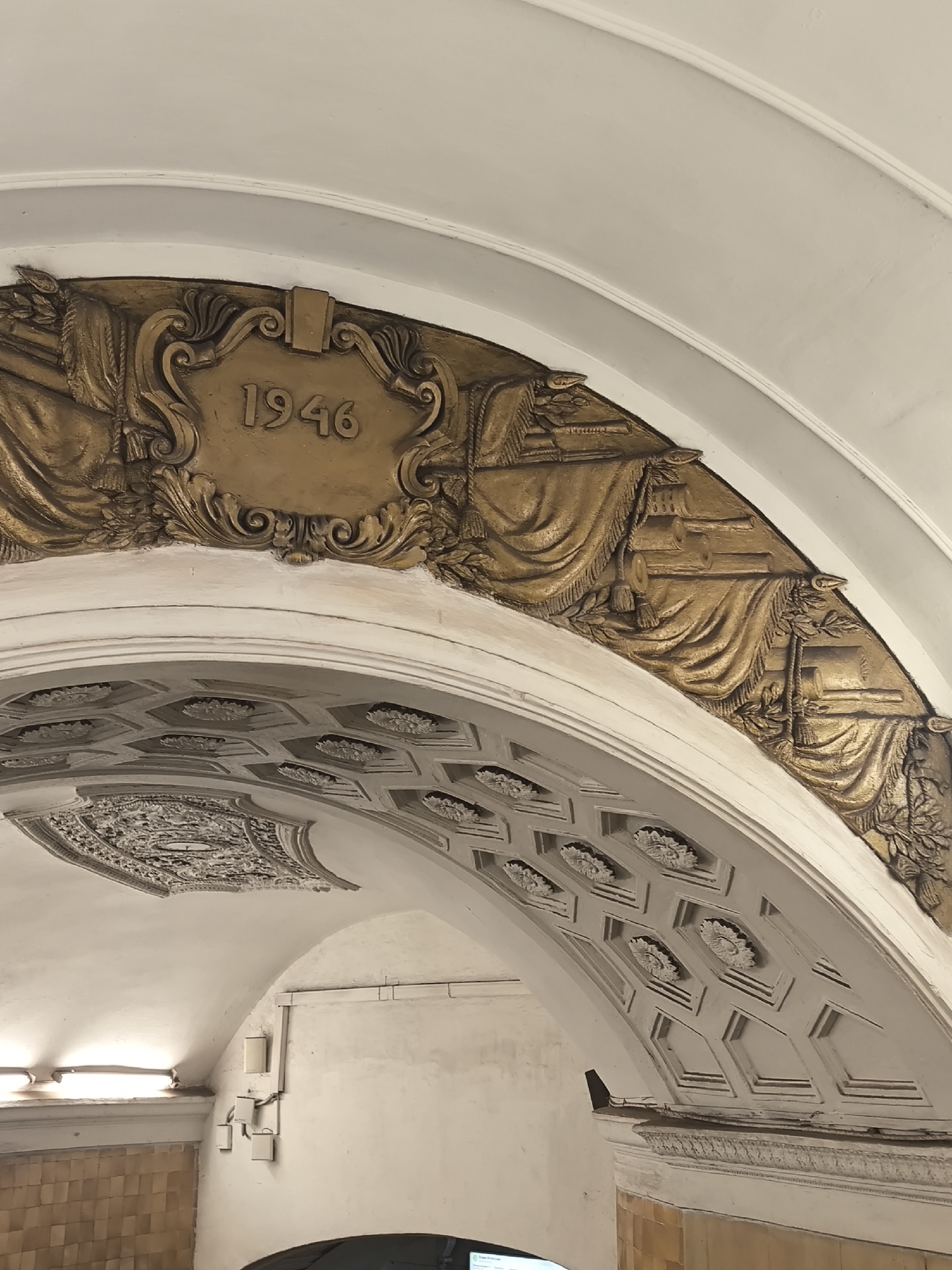 The image shows a section of an arched ceiling, possibly within a building. A prominent feature is a gold-colored, ornate relief displaying the number "1946". The relief is set against a white ceiling, which is detailed with geometric patterns and floral designs. The surrounding architecture appears to be old and possibly of historical significance. The location is listed as Unknown, Unknown.