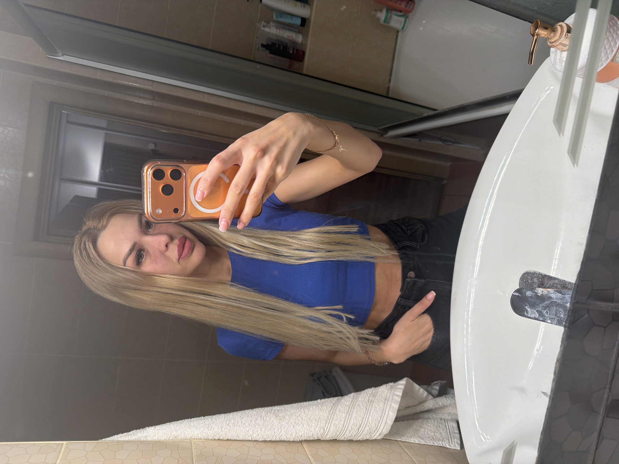 A woman with blonde hair is taking a mirror selfie in a bathroom. She is wearing a blue crop top and black jeans. She has her left hand in her pocket and is holding her phone with her right hand. The bathroom has beige tiled walls and a white sink. There is a towel hanging on the left side of the mirror. In the background, there is a shower with bottles of toiletries. The scene appears to be in a residential bathroom. There are no specific landmarks or features in the image to indicate the city of Kyiv, Ukraine.
