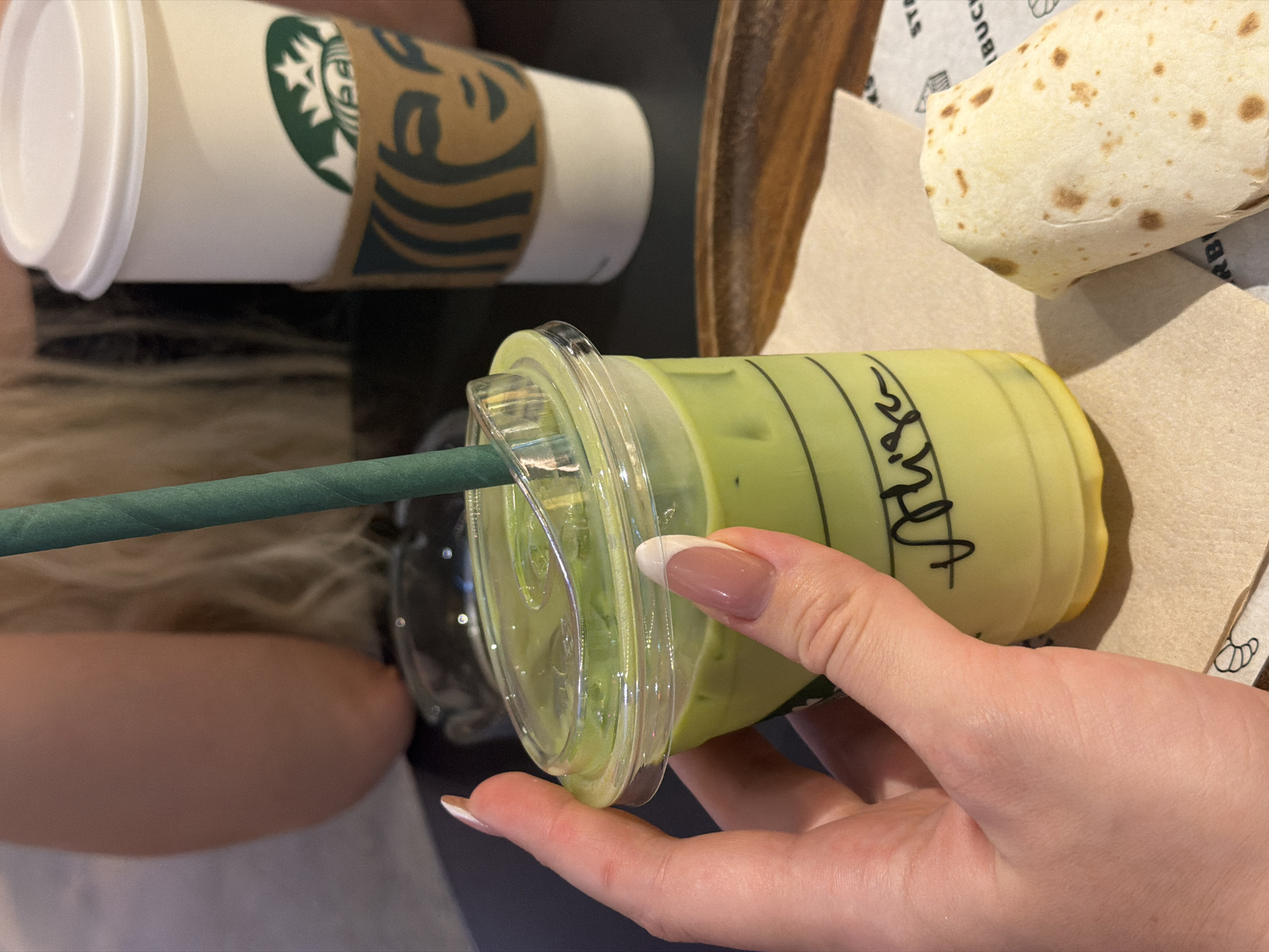 The image presents a close-up view of a meal, potentially at a Starbucks location. There is a Starbucks paper cup, with the logo visible. Adjacent to the cup is a partially visible food item, which appears to be a burrito or wrap. A hand is holding a plastic Starbucks cup containing a green liquid, likely a matcha latte. The cup has a green straw. The image gives the impression of a casual meal setting, probably in a café or restaurant. The specific location in Vracar Urban Municipality, Serbia cannot be determined from the image.