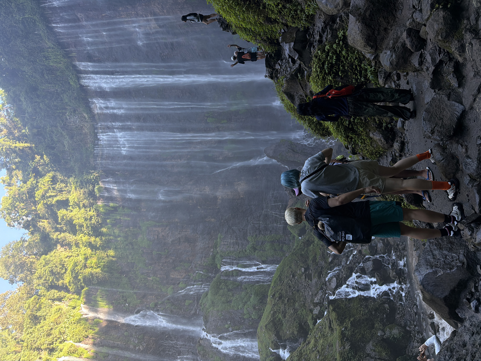 This image depicts the Tumpak Sewu waterfall in Sidomulyo, Indonesia. Multiple streams of water cascade down a sheer, moss-covered cliff face. Several individuals are present, either standing on a rocky path or on ledges near the waterfall. In the foreground, three people face away from the camera, looking towards the waterfall. One person on the right is standing on a higher ledge, looking out at the scene. Another group of two people can be seen further up the path, with their arms raised. The environment is lush and green, with dense vegetation covering the cliff and surrounding rocks. The lighting suggests it is daytime, possibly late afternoon, with the sun casting a warm glow on the upper foliage.