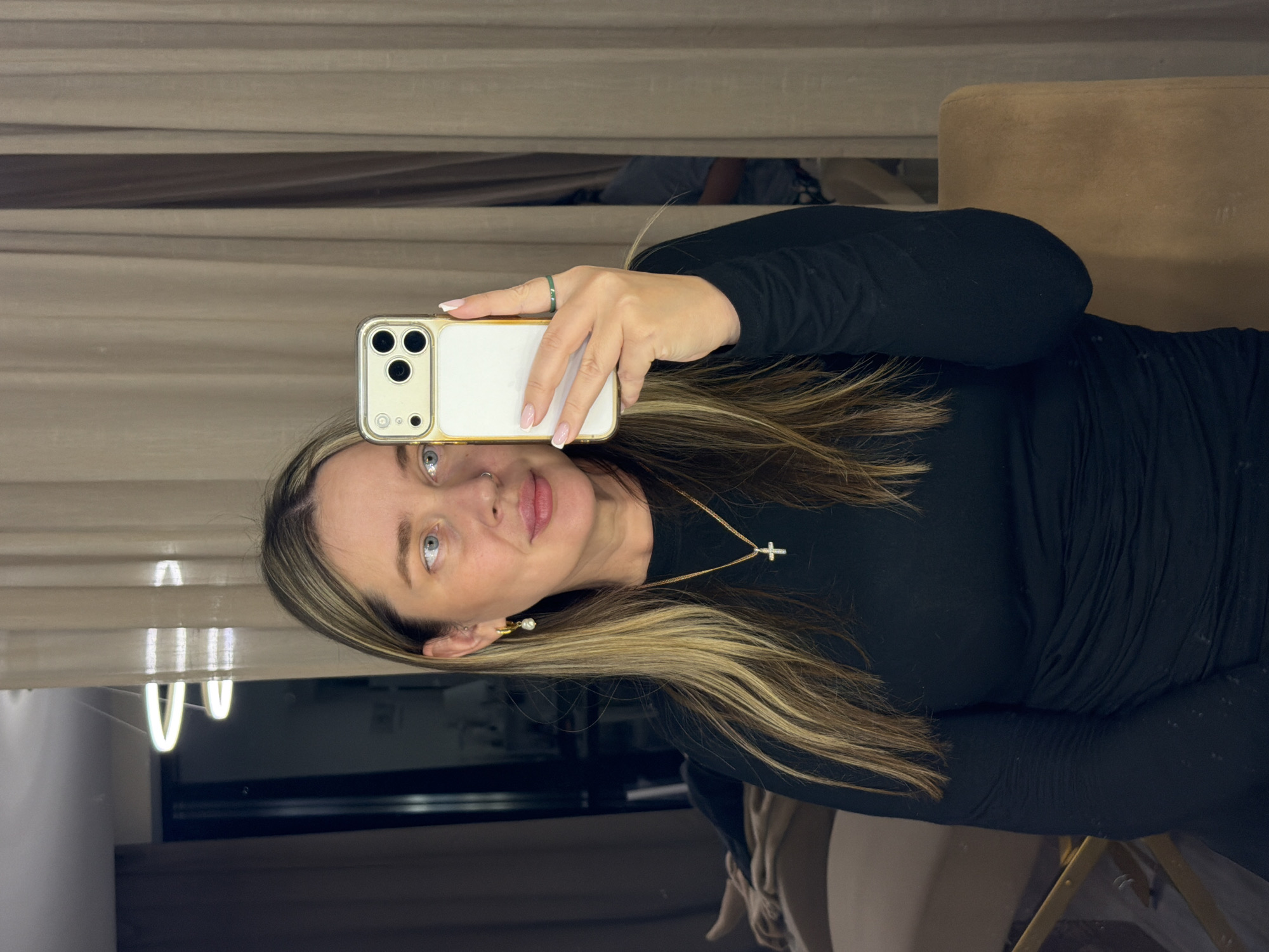A woman is taking a selfie with a white smartphone with a gold frame. She has blonde hair with dark roots and blue eyes. She is wearing a long-sleeved black dress and a gold necklace with a cross pendant. She also has an earring and a ring. The background appears to be a room with curtains, a table, and a chair. The location is Unknown, Unknown.