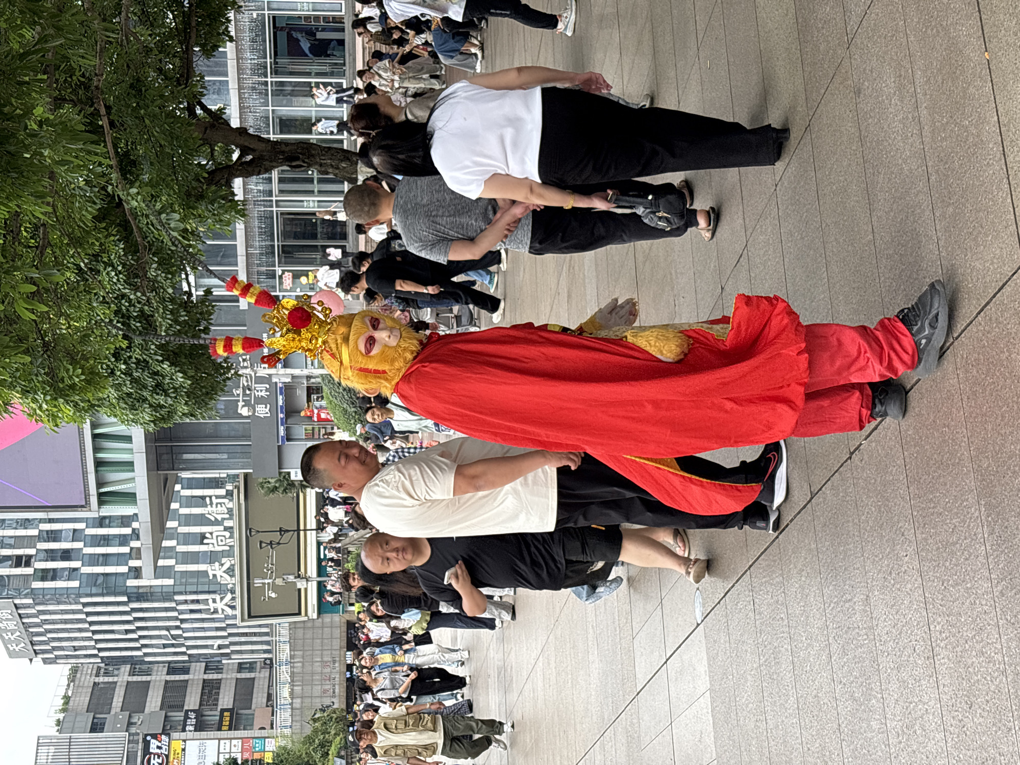 The image depicts a street performance of the Monkey King in Liangjiang, China. A person in a Monkey King costume, including a red cape, gold crown, and stylized mask, is supported by several people underneath the costume. The performance is taking place on a paved area lined with trees and modern buildings with signs in Chinese characters. A crowd of people is observing the performance.