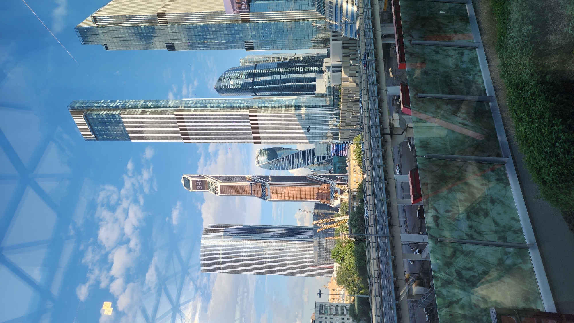 This image captures a view of the Moscow International Business Center, also known as Moscow City, on a clear day with a partly cloudy sky. The foreground features a reflective green glass barrier with a marbled pattern, possibly a sound barrier along a road. Behind this, an elevated highway is visible with several cars driving on it. In the background, a cluster of modern skyscrapers dominates the skyline.
The most prominent buildings are:
* A tall, slender skyscraper with a predominantly silver facade and dark horizontal banding, located on the far left.
* Another very tall, rectangular skyscraper with a silver-grey exterior and distinctive dark vertical elements, situated to the right of the first.
* A distinctive copper-colored skyscraper with an angular, tapering design, featuring the number "398" prominently displayed near its peak.
* A uniquely shaped, curved skyscraper with a blue glass exterior, resembling a twisted structure.
* A very tall, rectangular skyscraper with a light-colored facade and dark horizontal stripes, on the far right of the cluster.
Construction cranes are visible in the middle ground, suggesting ongoing development in the area. Residential buildings with a more traditional architectural style are located further in the distance, to the left of the skyscrapers. The overall scene depicts a modern urban landscape with a mix of towering commercial buildings and infrastructure.