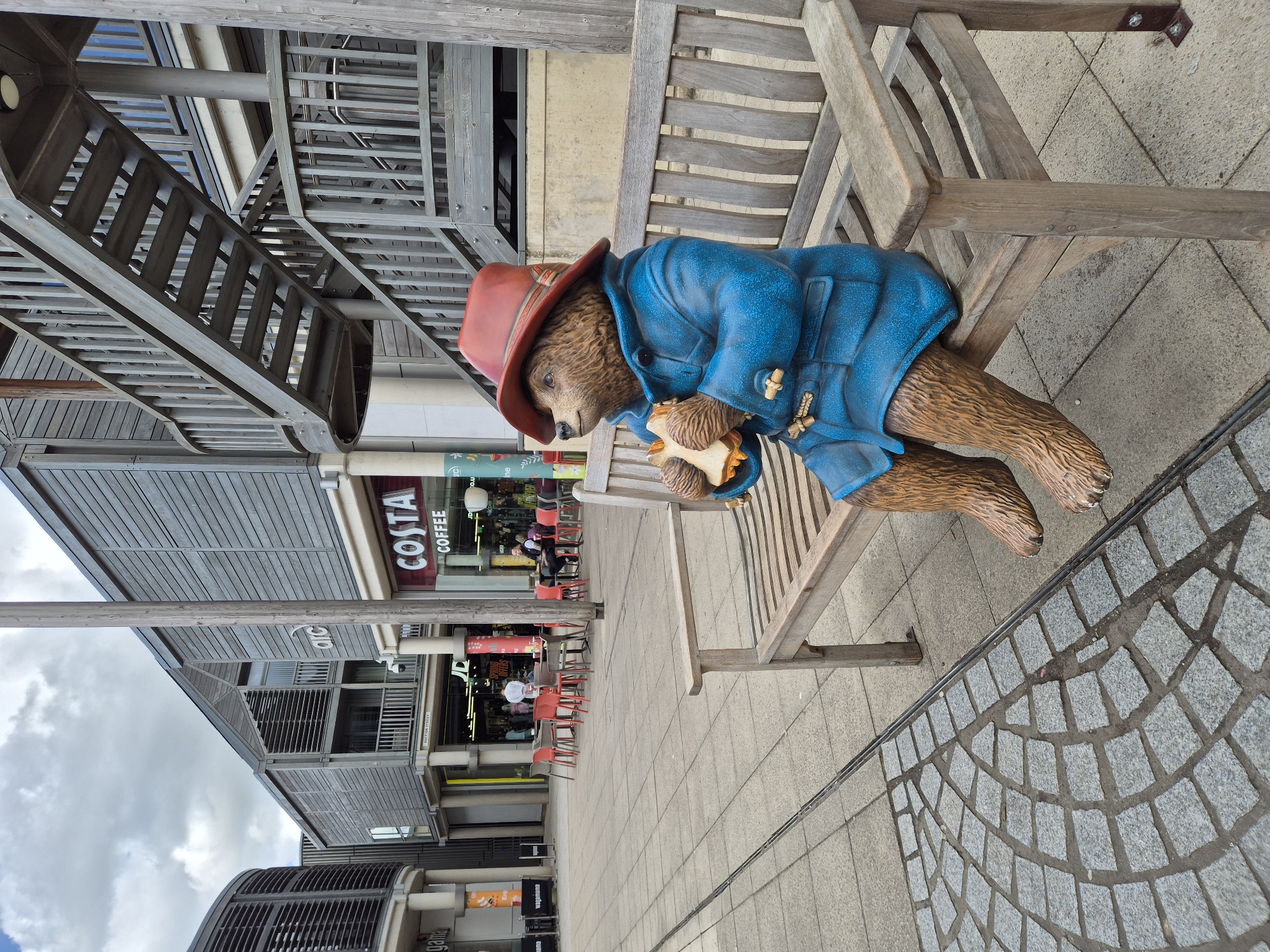 The image captures a statue of Paddington Bear seated on a wooden bench in a public area in West Suffolk, United Kingdom. Paddington wears his signature red hat and blue coat and holds a sandwich. Behind the bench, a modern building with metal stairs and grey cladding rises. A Costa Coffee shop is visible in the background, with people seated at tables outside. The ground is paved with a mix of grey stone and patterned tiles. The overall scene depicts a moment of whimsy within a contemporary urban setting.