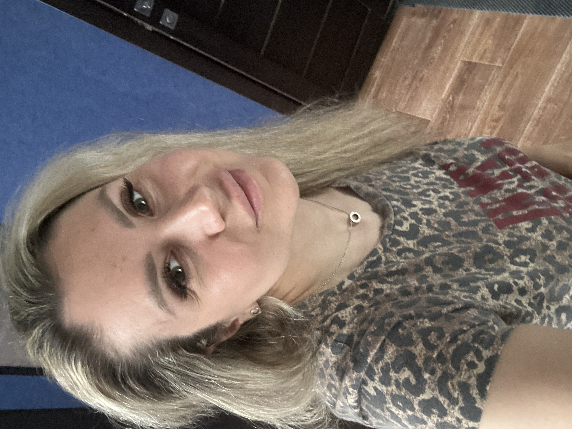 A woman with blonde hair is taking a selfie in an indoor setting. She is wearing a grey and black leopard print t-shirt with red lettering on the front and a silver necklace with a small circular pendant. She has light-colored earrings and her makeup includes mascara and lip gloss. The background is a blue wall and a wooden floor, with a dark brown door visible on the right side of the frame. The image was taken in Novoaltaysk, Russia.