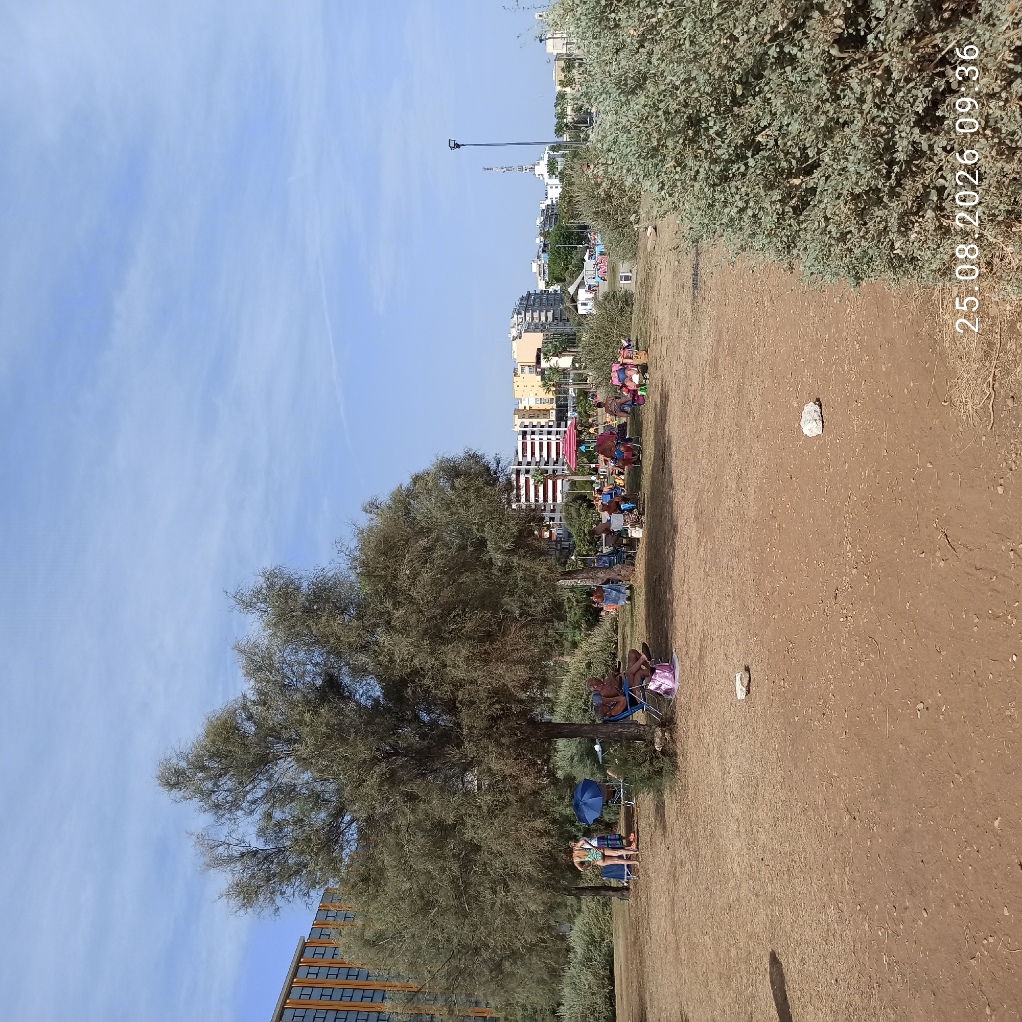 The image depicts a park-like area with a sandy ground, sparse vegetation, and a few trees, under a clear blue sky. In the background, there is a cityscape with several multi-story buildings of varying architectural styles, some appearing to be residential or hotels. A tall communication tower is visible amongst the buildings.
The foreground shows several individuals and groups enjoying a leisurely day outdoors. On the right side of the image, two people are seated in blue lounge chairs, with one of them appearing to be reading. A pink tote bag is on the ground next to them. To the left of these individuals, further into the scene, a group of people are gathered, some seated in chairs and others standing. One person is wearing a patterned swimsuit. A blue umbrella is set up, and there is a pink and white striped umbrella further back. Some people are shaded by trees. The overall atmosphere is relaxed, suggesting a picnic or a casual outing in a public park.
The scene is likely located near the coast, given the sandy ground and the presence of beach umbrellas. The date and time stamp in the lower right corner indicates "25.08.2026 09:36".
While the prompt mentions the city of Bari, Italy, there are no specific visual cues within the image that definitively confirm this location without external information. The architecture of the buildings and the style of the communication tower could be consistent with many coastal European cities.