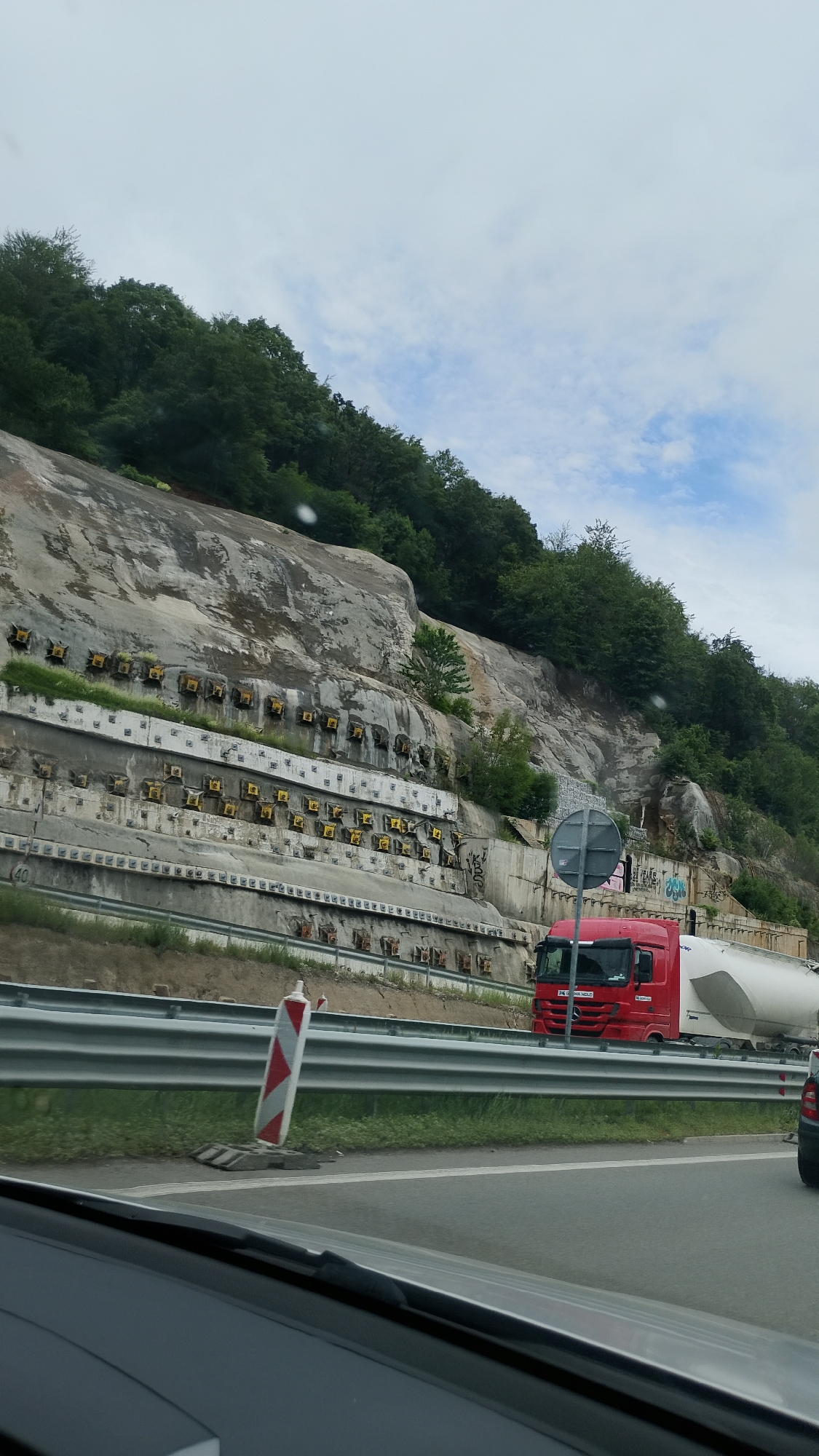 The image depicts a highway scene in Vrachesh, Bulgaria. On the right side of the road, a red semi-trailer truck with a white tanker trailer is traveling. The truck has a license plate that is partially visible, showing "JA JIMA HOLM". In the background, a steep, rocky mountainside is visible, reinforced with multiple levels of concrete structures and yellow anchors. The mountainside is covered with trees. A guardrail separates the road from the embankment. A traffic cone with red and white stripes stands on the side of the road. The sky is overcast with some clouds. The scene appears to be a construction or maintenance area along a mountain pass.