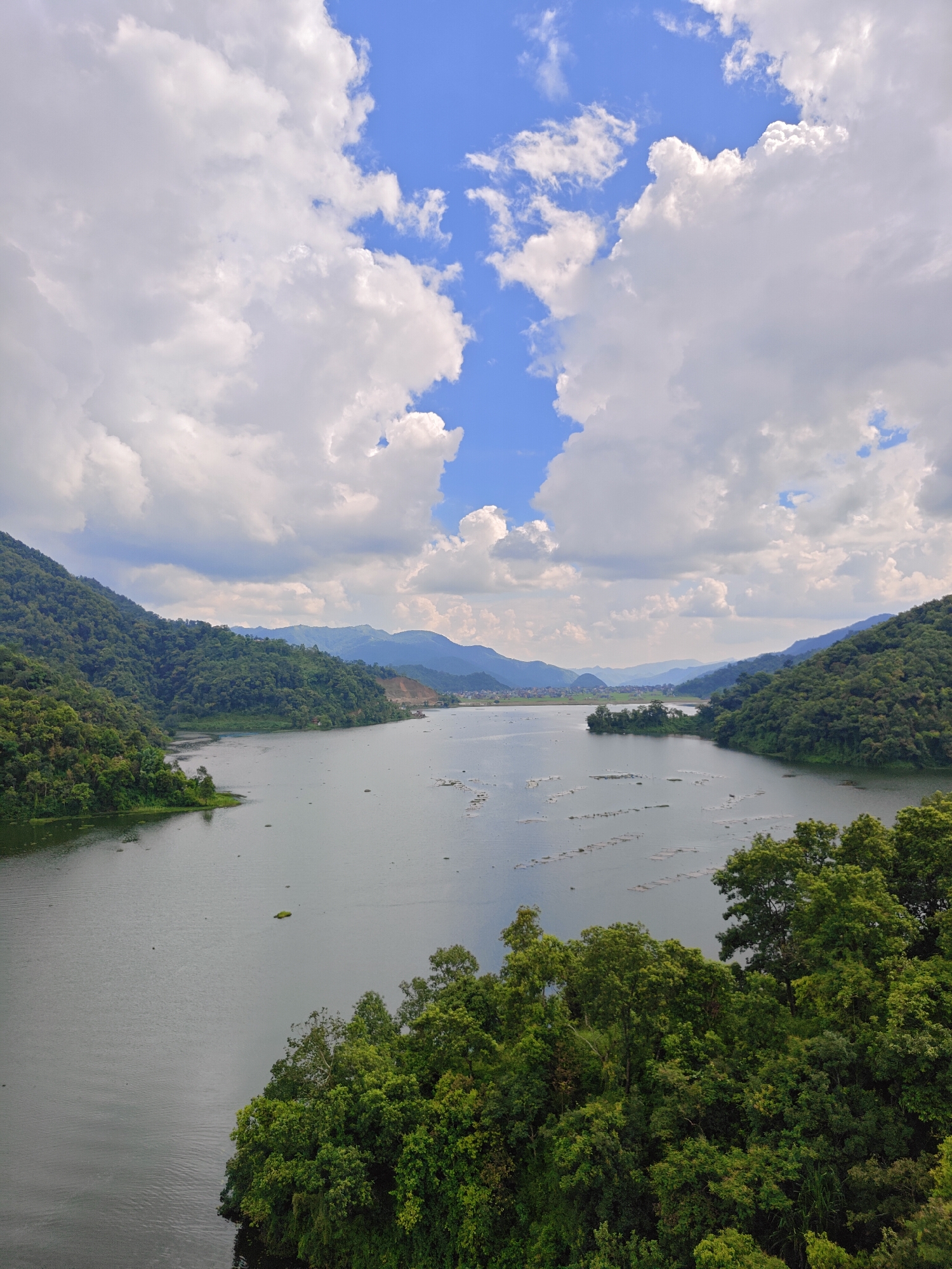 The image shows a wide, tranquil lake surrounded by lush green hills and mountains. The sky is a vibrant blue, dotted with fluffy white clouds. In the foreground, the dense foliage of trees frames the bottom of the scene, creating a sense of depth. The water surface reflects the sky and the surrounding landscape, with subtle ripples indicating a gentle breeze. In the distance, the hills recede, becoming softer and more muted in color, eventually blending into the hazy outlines of mountains on the horizon. A few scattered structures and possibly aquaculture farms are visible on the water. This is likely Begnas Lake, a prominent natural attraction in Lekhnath, Nepal. The scene is peaceful and evokes a sense of natural beauty and serenity.