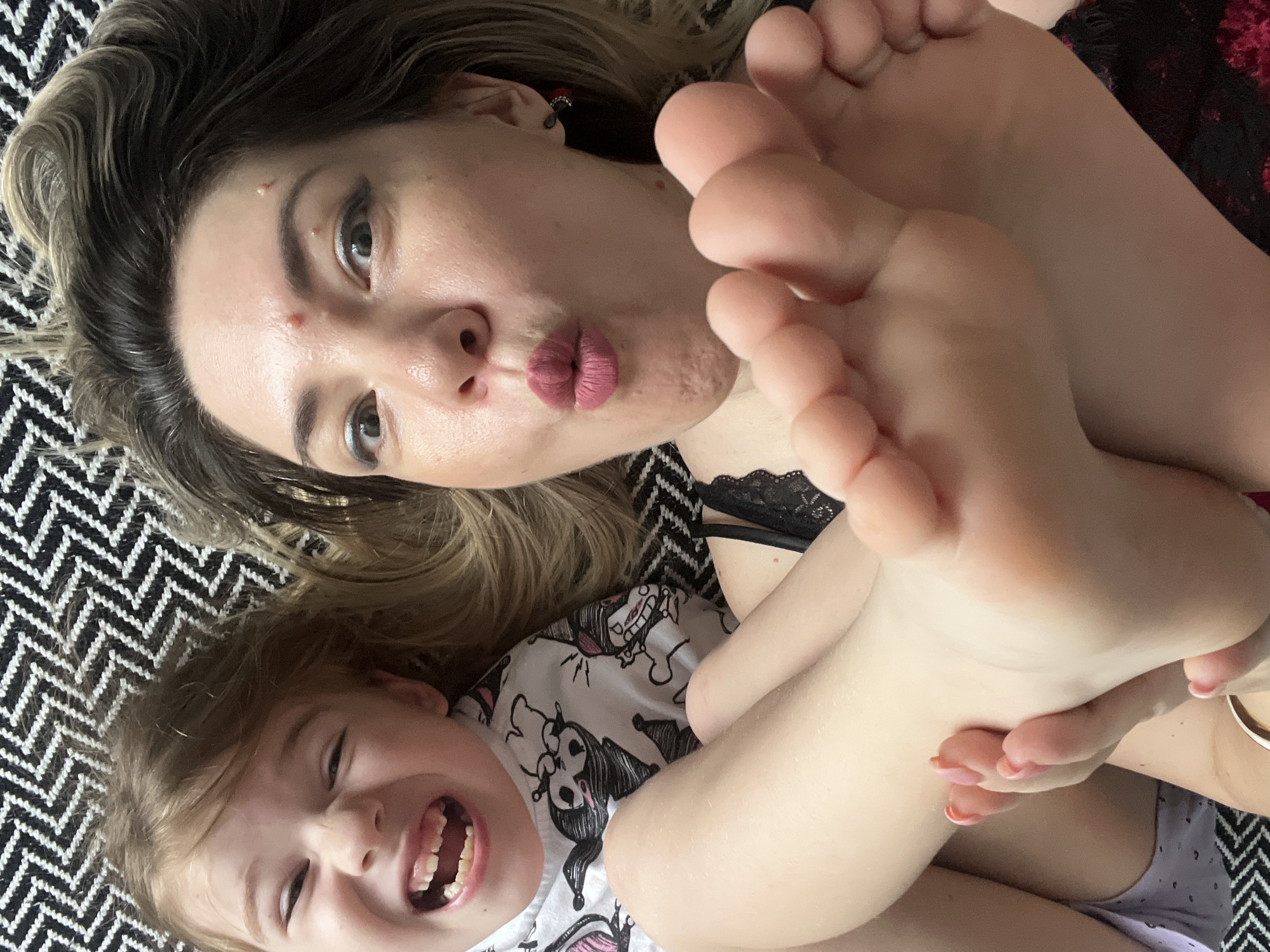 The image shows a close-up, high-angle shot of a woman and a child lying down on a patterned surface. The woman, in the foreground, is making a kissing face, with her lips puckered. Her feet are prominently displayed, with one foot held up towards the camera. She is wearing black lace lingerie. The child, to the left and slightly behind the woman, has their mouth wide open in a joyful expression, with their teeth visible. The child is wearing a white t-shirt with black cartoon characters printed on it. The background is a black and white zigzag patterned surface, possibly a blanket or rug. The lighting is bright, casting some shadows. The image appears to be a selfie taken by the woman. The location of this scene is unknown, and there is no information to suggest it is in Sochi, Russia.