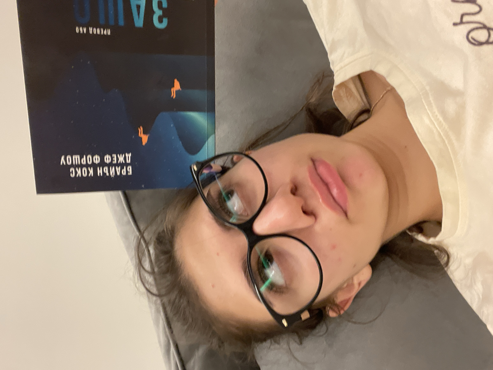 A woman is lying on a gray couch, holding a book over her head. The book cover features a dark blue background with stylized illustrations of mountains and small orange figures. The title, likely in a non-English language, is printed in a large, teal font, and the authors' names are below in a smaller font. The woman is wearing black-rimmed glasses and a light-colored shirt. The location is not discernible from the image.