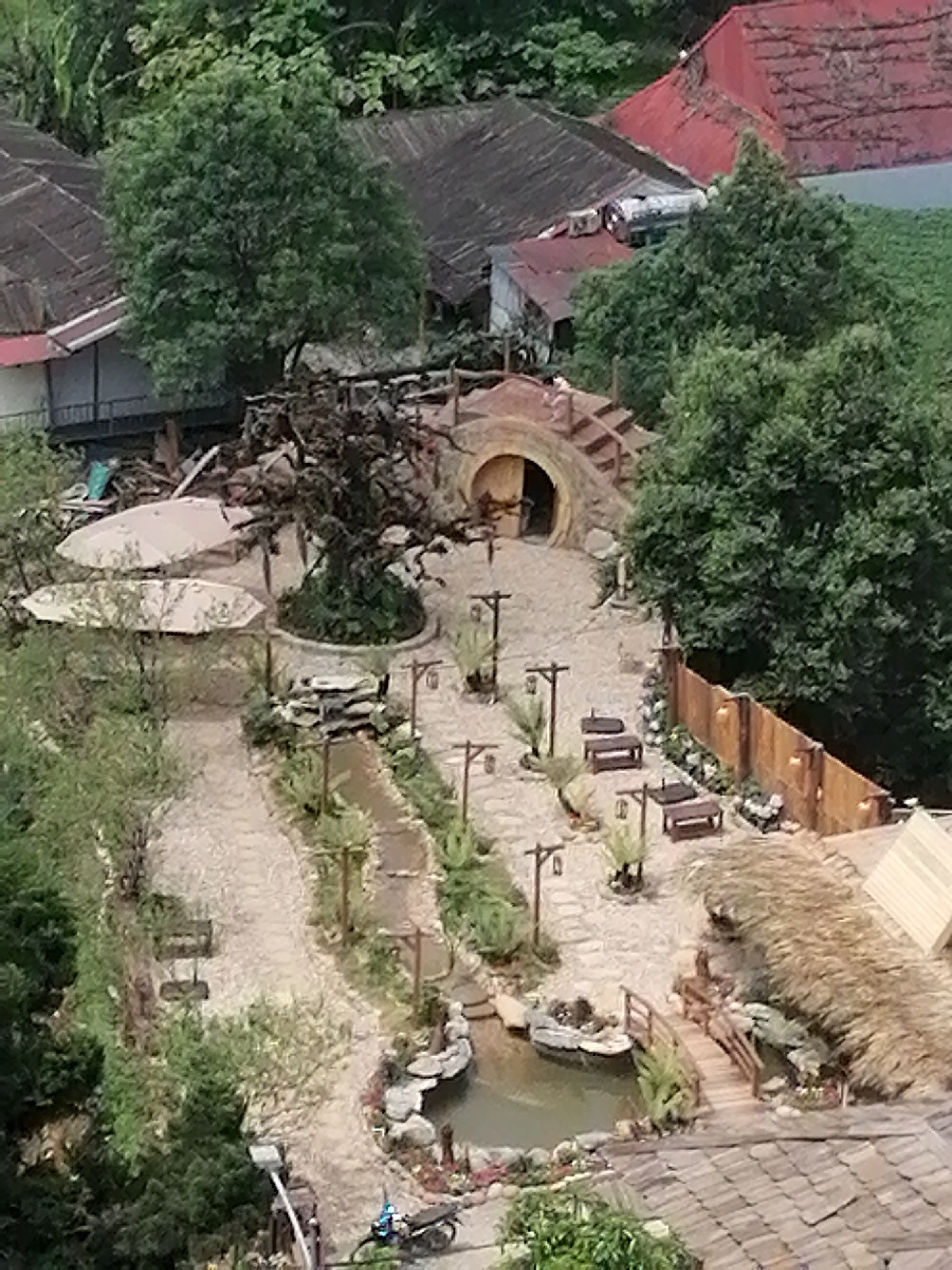 The image shows an elevated view of a landscaped area in Sa Pa Ward, Vietnam. A pathway extends through the scene, leading to a hobbit-hole structure with a rounded entrance and stairs climbing its exterior. Several seating areas with benches are arranged along the pathway. A small pond and a thatched-roof structure are visible towards the lower right. Dense foliage and buildings with red and grey roofs surround the landscaped area. A parked motorbike is visible near the pond. Parasols are used to create seating in a more open area along the upper left side of the image. The overall setting appears to be a tourist attraction.