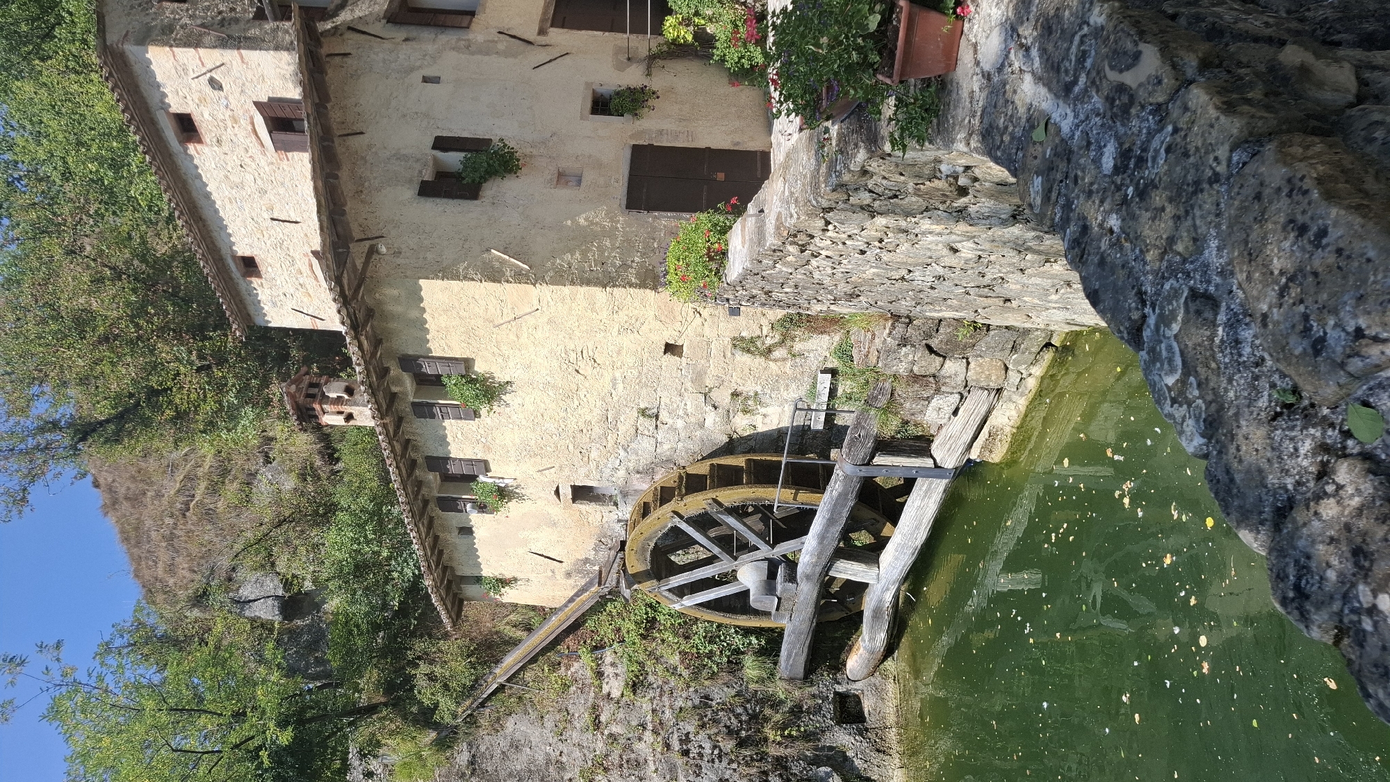 The image depicts a historic watermill situated in a lush, natural setting. The mill is constructed of stone and plaster, with a prominent wooden waterwheel. The waterwheel is partially submerged in a body of green water, which appears to be a pond or canal. Wooden planks are visible in and around the water, forming a walkway or support structure for the mill.

The mill building is two stories tall and has a tiled roof. Several windows with wooden shutters are visible on the facade. A brick chimney rises from the roof. In front of the building, a stone wall runs along the edge of the water, and potted plants with red and pink flowers add a touch of color.

The background features a steep, vegetated hillside with rocky outcrops and trees. The sky is clear and blue. The overall impression is one of rustic charm and historical significance.

This watermill is located in the municipality of Pieve di Soligo, Italy, in the Veneto region.