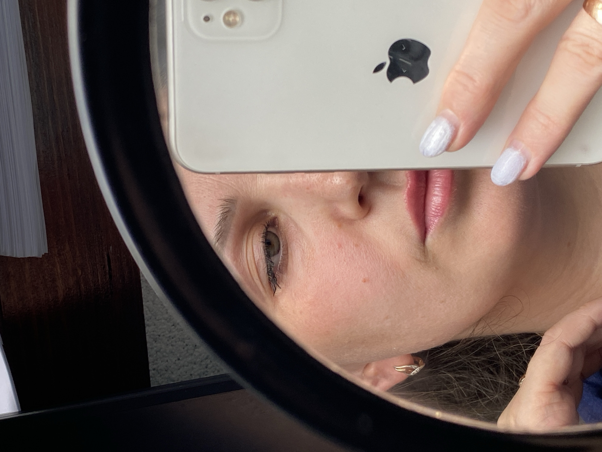A woman is taking a selfie in a mirror, holding an iPhone 11 or similar model, with a white case. The phone covers part of her face, obscuring her forehead and chin. Her nose and lips are visible between the phone and her fingers. The woman has fair skin, an eye with visible eyelashes, and pink lips. She has silver nail polish on her fingernails. She is wearing a gold ring and earrings. Part of her hair is visible near her ear. The mirror is circular with a thick black frame. The background contains partially visible sheets of paper and a wooden surface. No specific location details for Markovskii, Russia are apparent in the image.