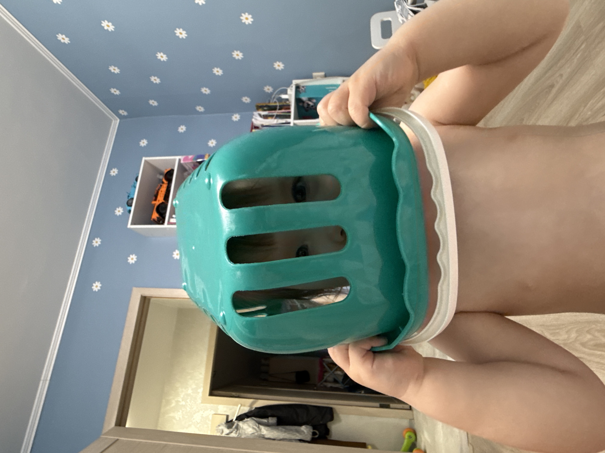 A close-up, tilted shot depicts a child with fair skin and dark hair holding a turquoise plastic container over their head, so their eyes are visible through the holes. The background shows a room with light blue walls adorned with small white flower patterns. To the left is a grey wall section, and a white shelf unit containing toy cars is also visible. A doorway with a light-colored frame is in the lower left, leading to a glimpse of a room with clothes and other items. A wooden floor is partially seen on the right.