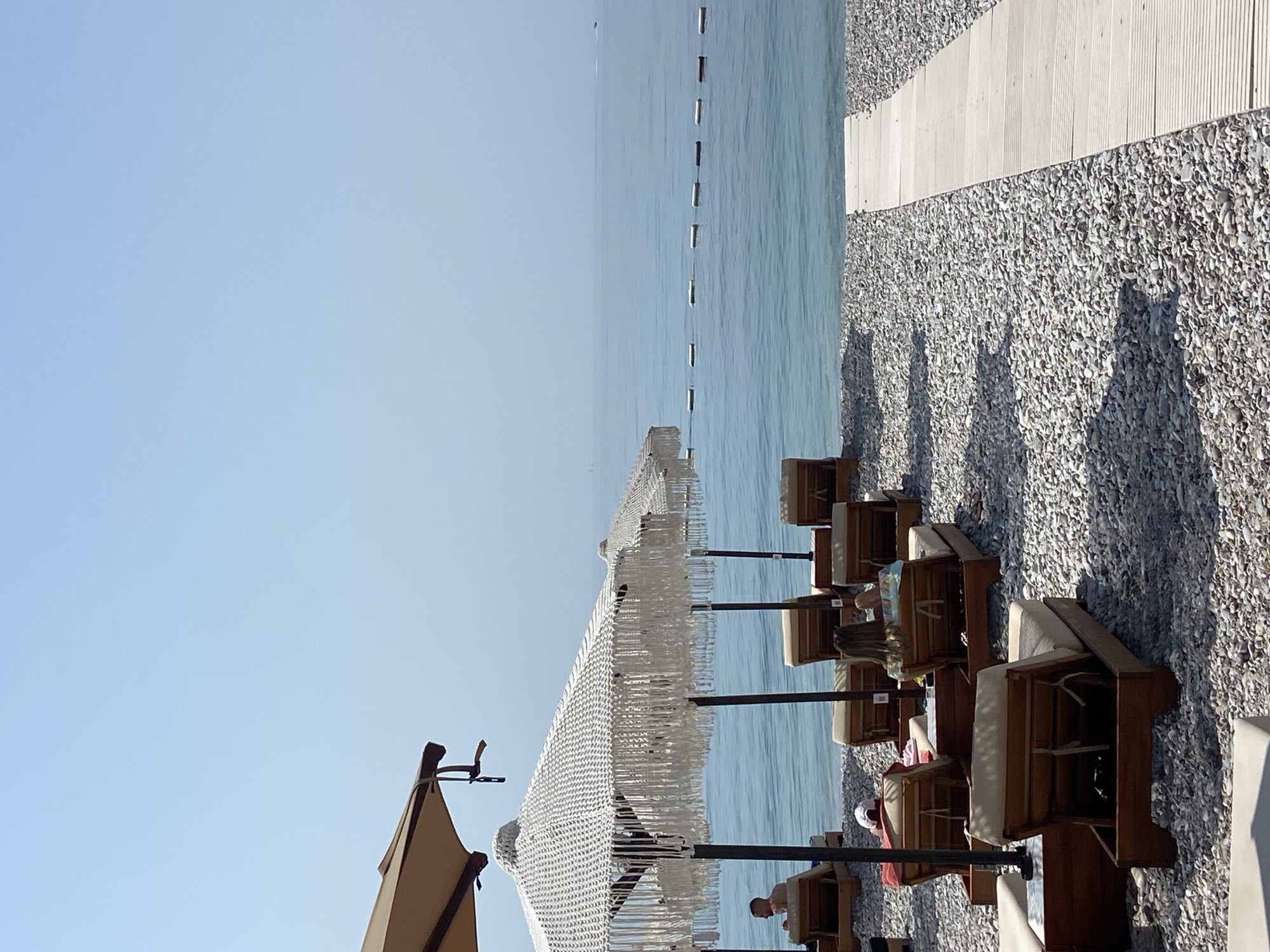 This is a photograph of a beach scene in Prijevor, Montenegro. The image captures a section of a pebble beach with several lounge chairs and umbrellas set up for patrons. The sea is calm and blue, stretching out to the horizon under a clear sky. A wooden walkway is visible on the right side of the frame, leading towards the water. Several people are seated on lounge chairs, relaxing under the shade of umbrellas, suggesting a leisurely atmosphere. One person is visible in the water near a distant boat. The scene is well-lit, indicating it was taken during daylight hours.
