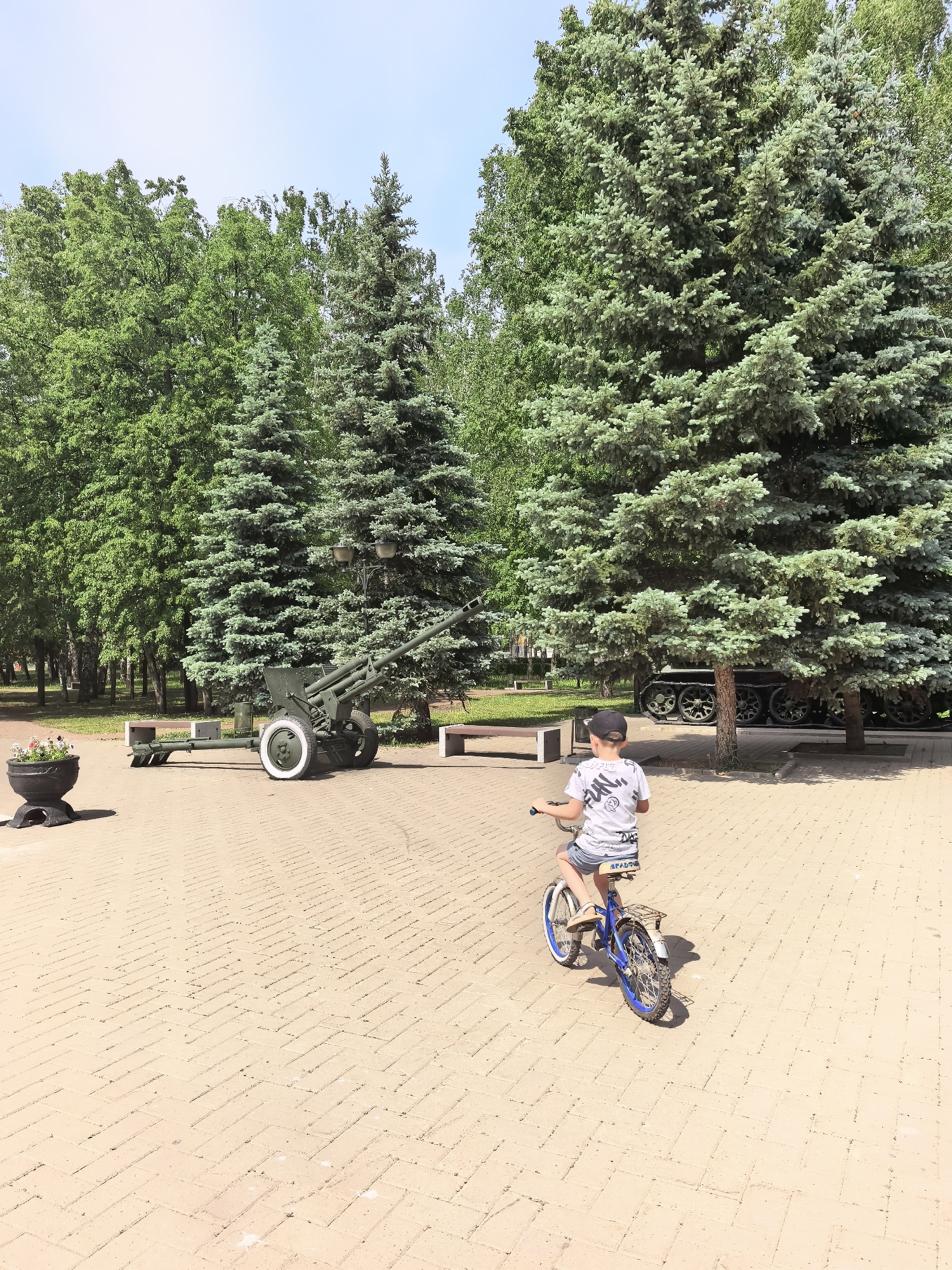 A young boy rides a blue bicycle on a paved brick surface in a park-like setting. The brick pavement extends into the distance, with a pattern of laid bricks visible. The boy is wearing a white t-shirt with black lettering, denim shorts, and a baseball cap. Behind the boy is a military cannon with white tires, positioned on a concrete base. Various trees, including evergreen and deciduous types, are scattered throughout the scene. To the right of the cannon, the tracks of a tank are visible. A stone flower pot stands to the left of the frame. The setting appears to be a war memorial located in the city of Sterlitamak, Russia.