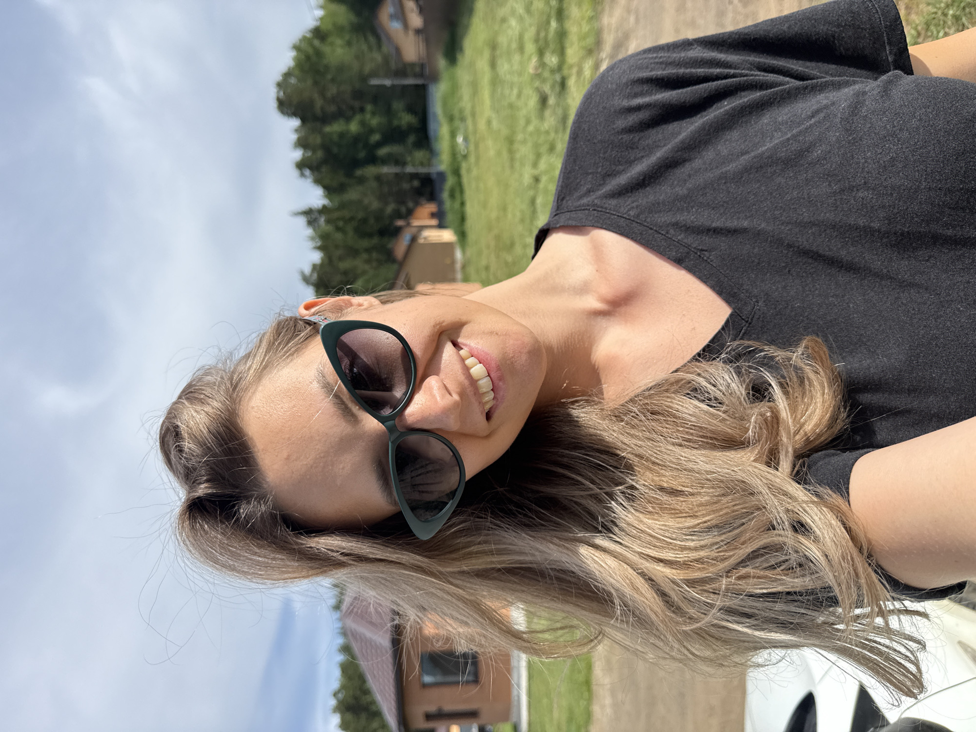 A woman with blonde hair, wearing a black V-neck t-shirt and dark green cat-eye sunglasses, smiles at the camera. Her hair is styled loosely with some strands falling over her face and shoulder. The background shows a rural setting with a house under construction or renovation, a grassy field, and trees. The scene appears to be in Chaikovsky, Russia.