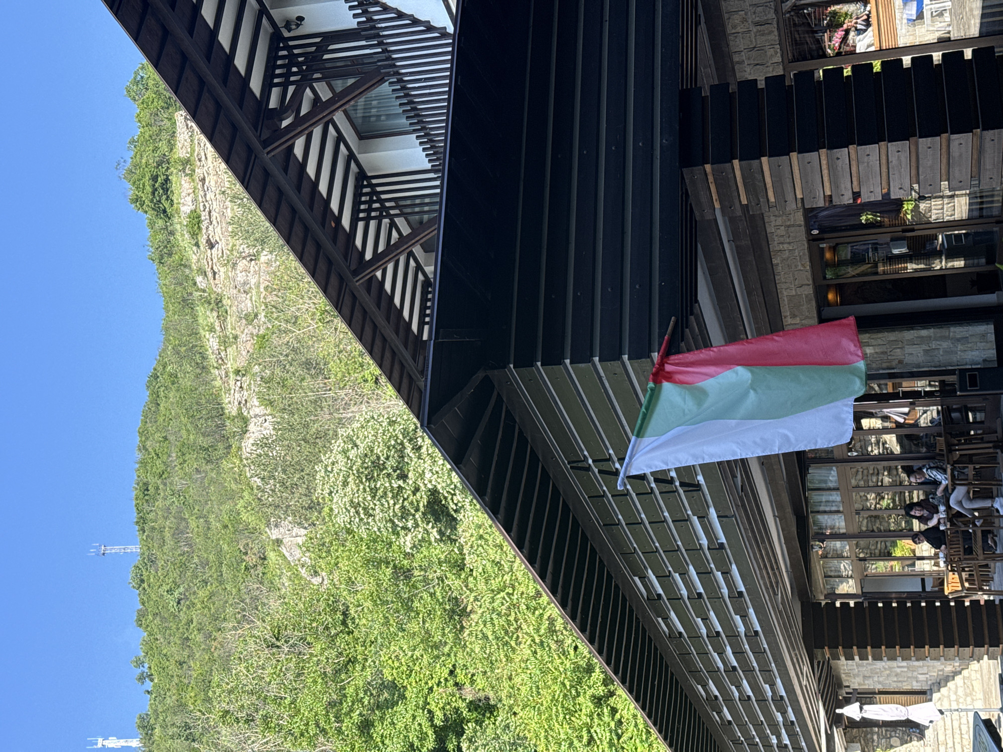 The image shows an outdoor scene near a building in Veliko Tarnovo, Bulgaria. A Bulgarian flag hangs from the building's exterior. In the background, there is a steep, green, tree-covered hill and a clear blue sky. A small transmission tower can be seen near the top of the hill. The building features a modern, possibly cantilevered design with dark wood or dark-painted slats and a lighter, natural wood color on the structure. There are people seated at tables outside the building. The scene appears to be during the daytime with strong sunlight.