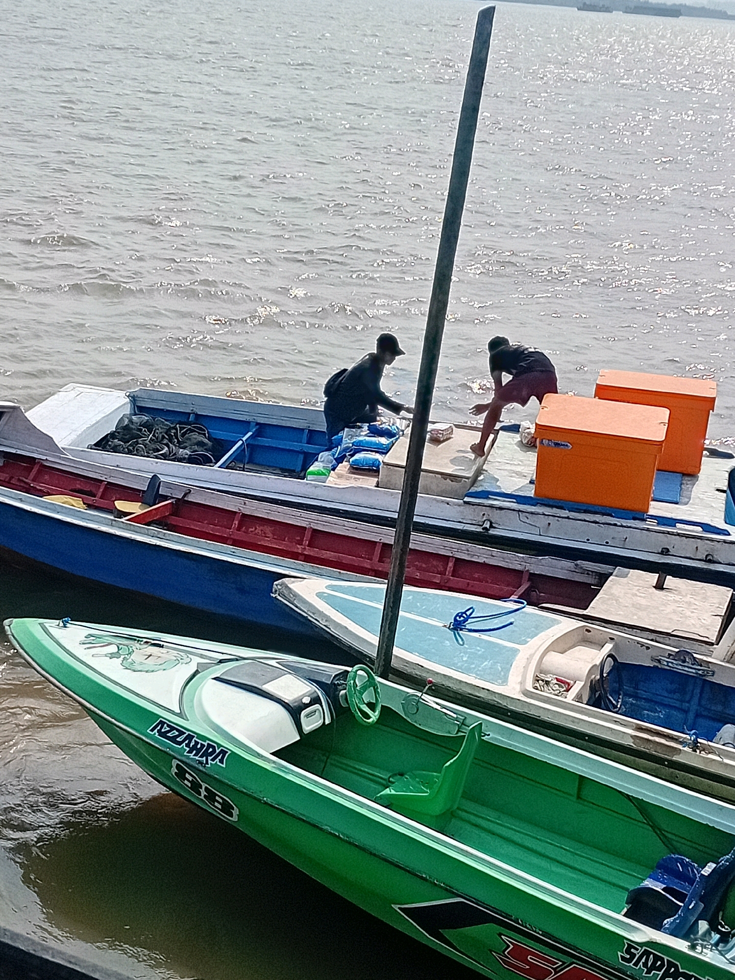 Two individuals, appearing to be fishermen, are engaged in activities on boats moored in the water. The fisherman on the left, wearing a dark long-sleeved shirt and a baseball cap, is facing away from the camera and appears to be handling fishing gear or supplies within a boat. The fisherman on the right, wearing a dark t-shirt and maroon shorts, is bent over, reaching towards or interacting with items on the deck of a larger boat.
The boats are docked together. The foreground features a bright green, sleek speedboat with "NZARRA BB" emblazoned on its side. Behind this, a larger, older-style boat is visible, painted with a blue hull and a red interior. This boat contains several orange storage containers. Further back, another boat with a white exterior and a blue interior is partially visible. A tall, thin pole, possibly part of a mooring or antenna, stands vertically in the center of the image, casting a shadow.
The water is calm with a shimmering surface, reflecting the bright sunlight. The scene appears to be a waterfront area, likely a harbor or dock, in Sungai Meriam, Indonesia. The overall atmosphere suggests routine activity related to fishing or boat maintenance.